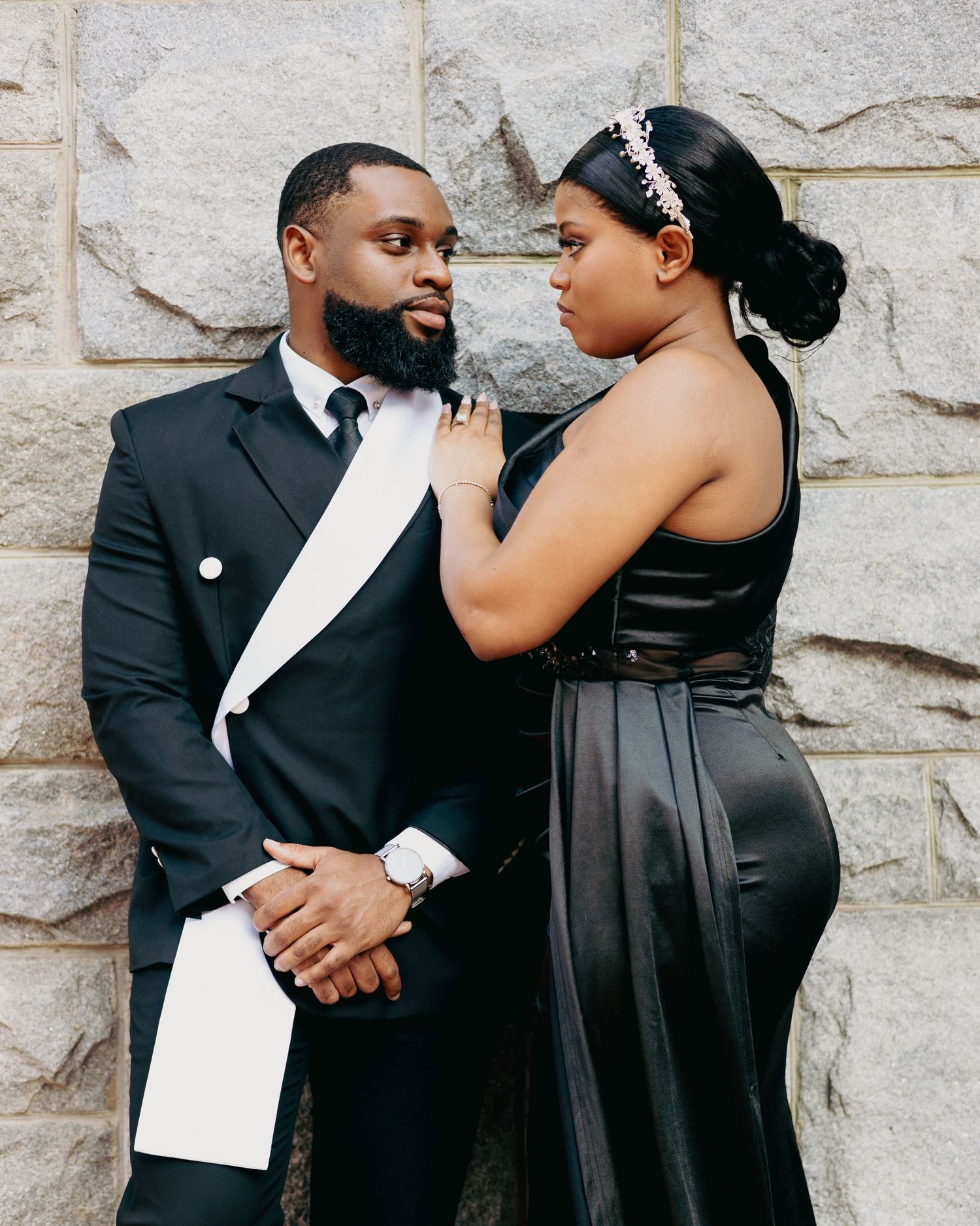 A couple in elegant black formal wear poses romantically against a stone wall, the woman adjusting the mans bow tie.