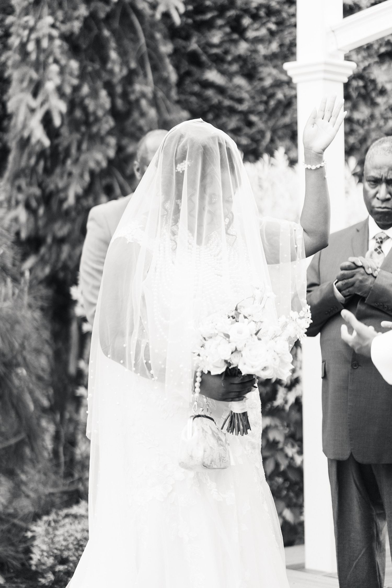 Black and white photo of veiled bride holding white bouquet at outdoor wedding altar.