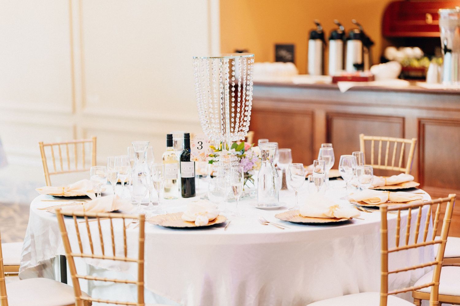 Elegant wedding reception table with gold Chiavari chairs, white linen, crystal glasses, and wine bottles in a bright venue.