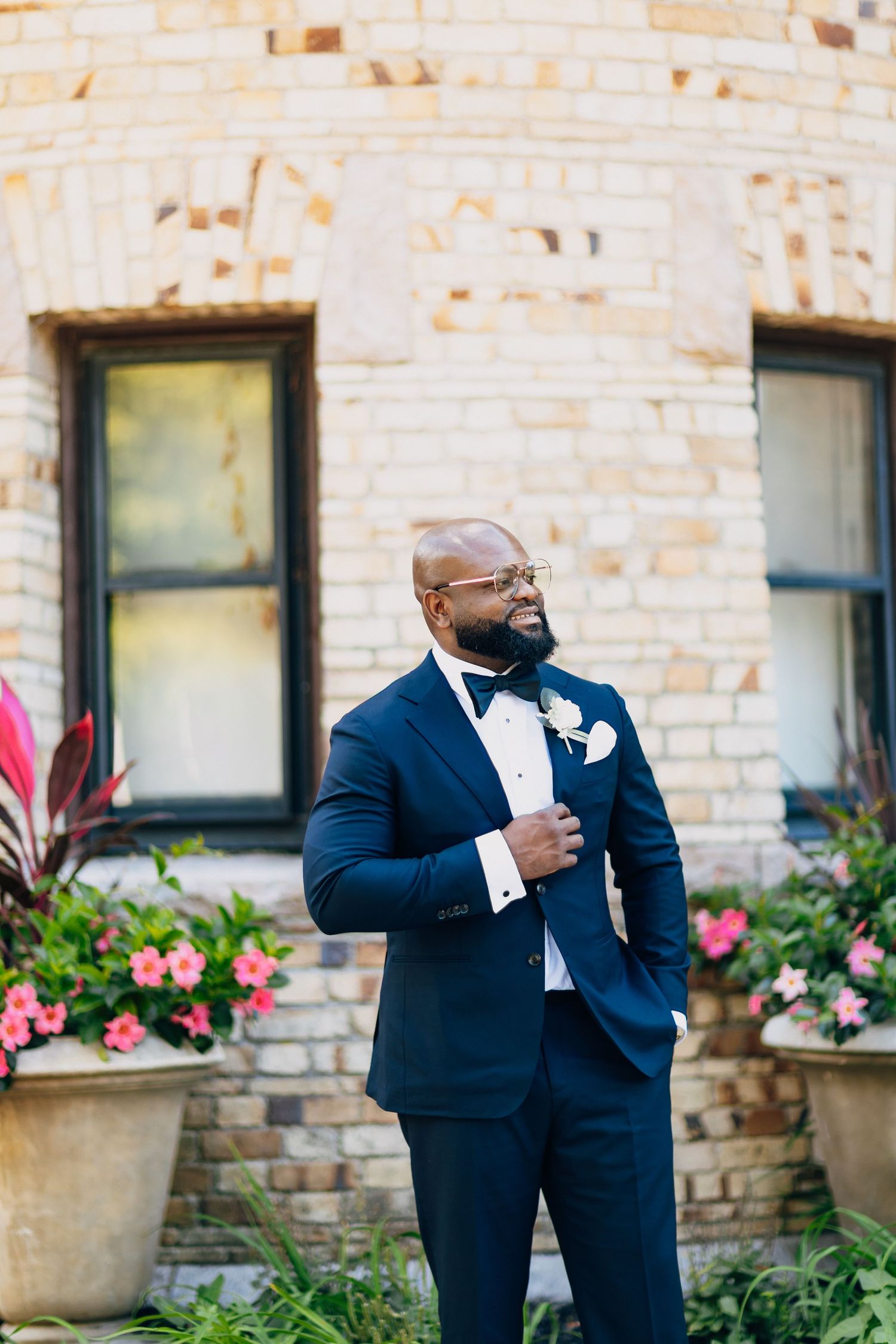 Dapper groom in navy tuxedo with bow tie and boutonniere posing outside brick building.