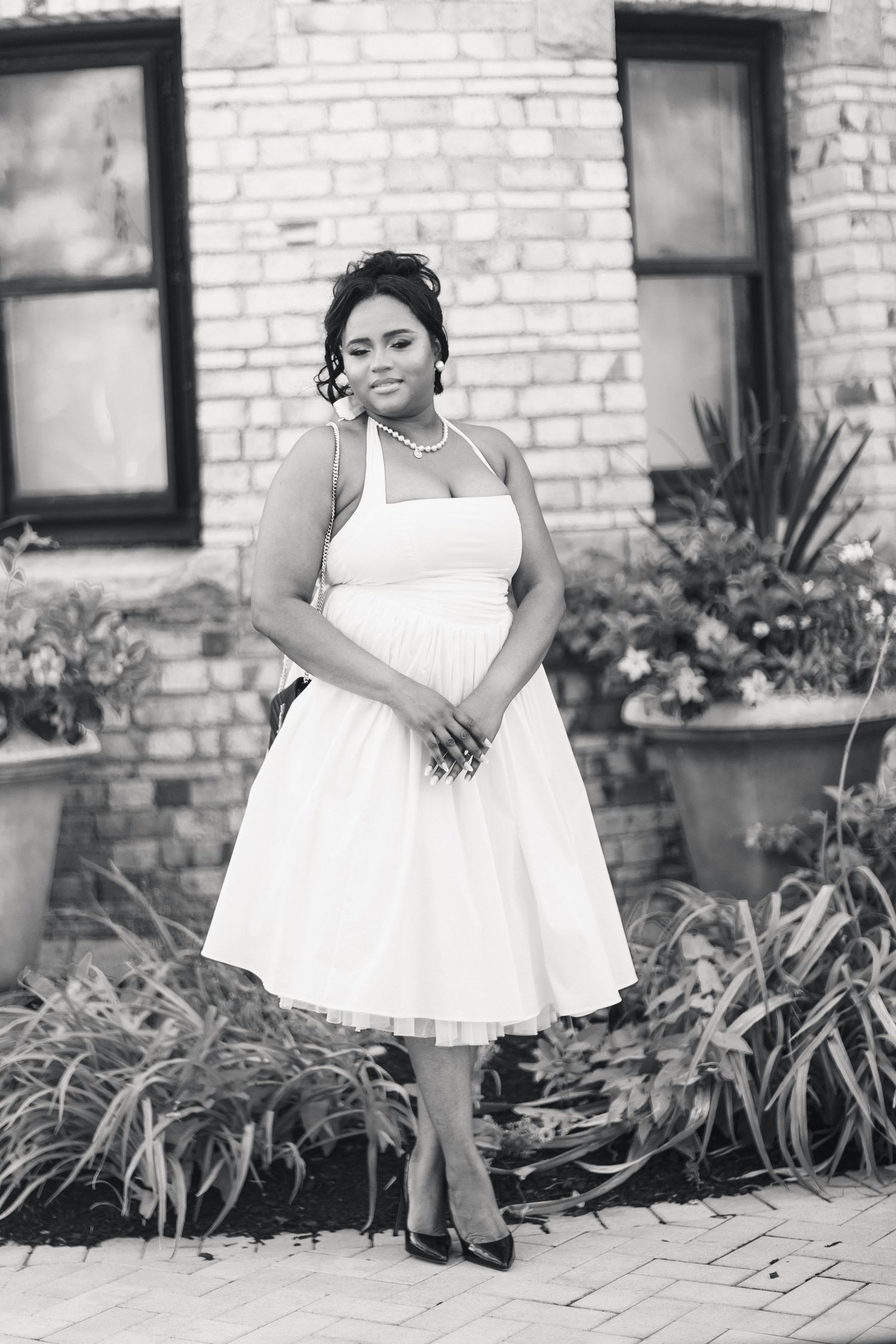 Bride poses in vintage-style white halter tea-length dress with pearls before brick building in black and white.