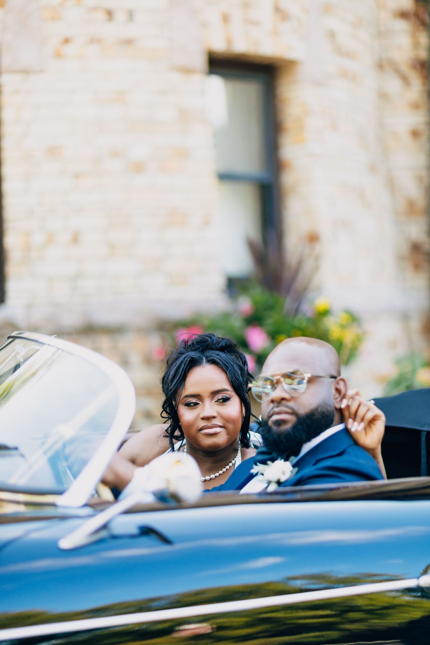 A bride and groom in formal wedding attire sit together in a black convertible car outside a stone building.