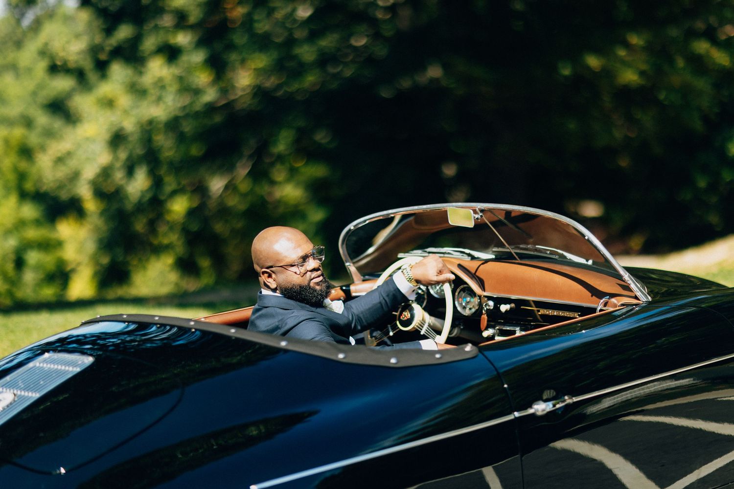 Bald groom in navy suit driving a classic black vintage convertible sports car.