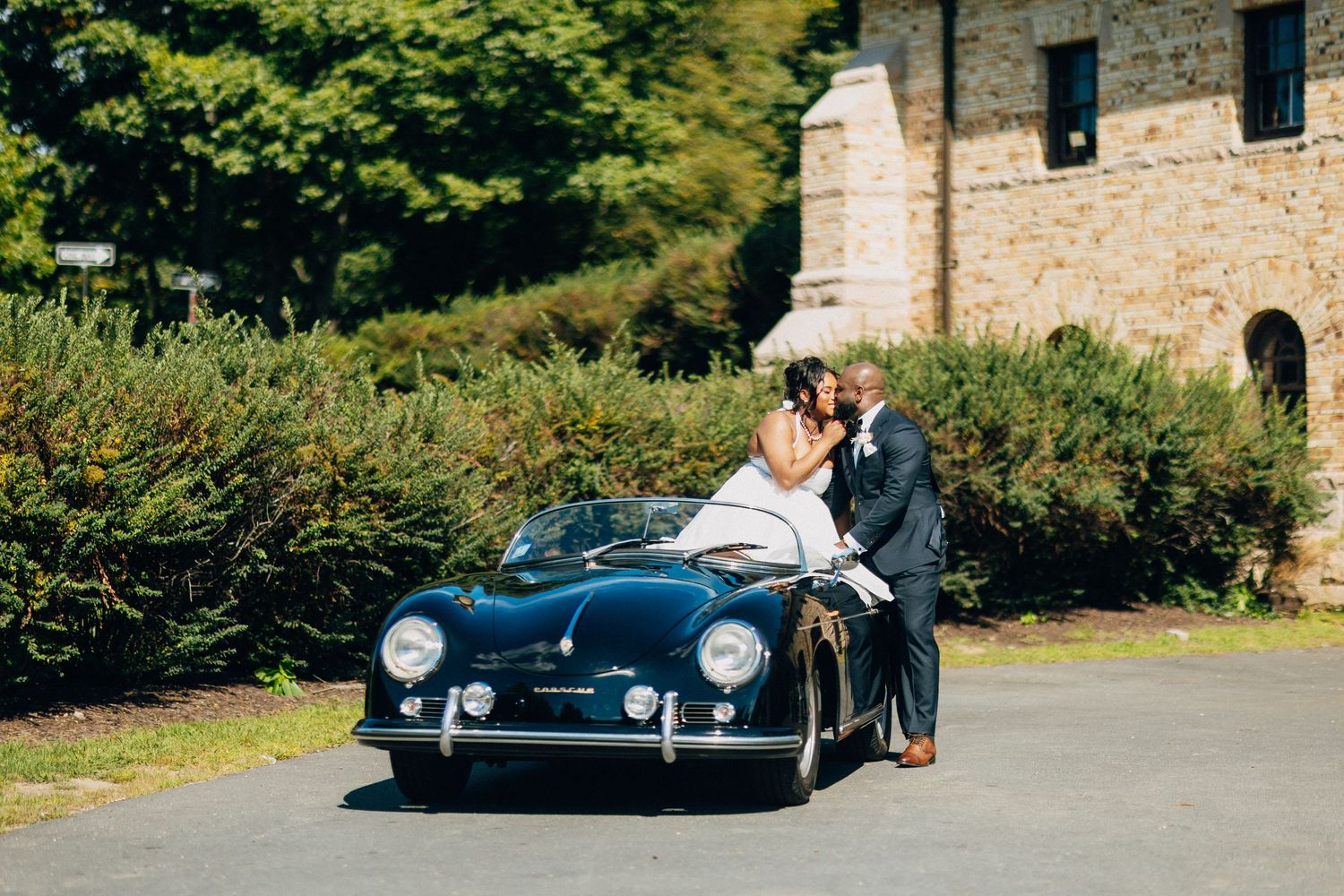 Bride and groom sharing a romantic moment leaning on a classic black Porsche 356 near stone venue.