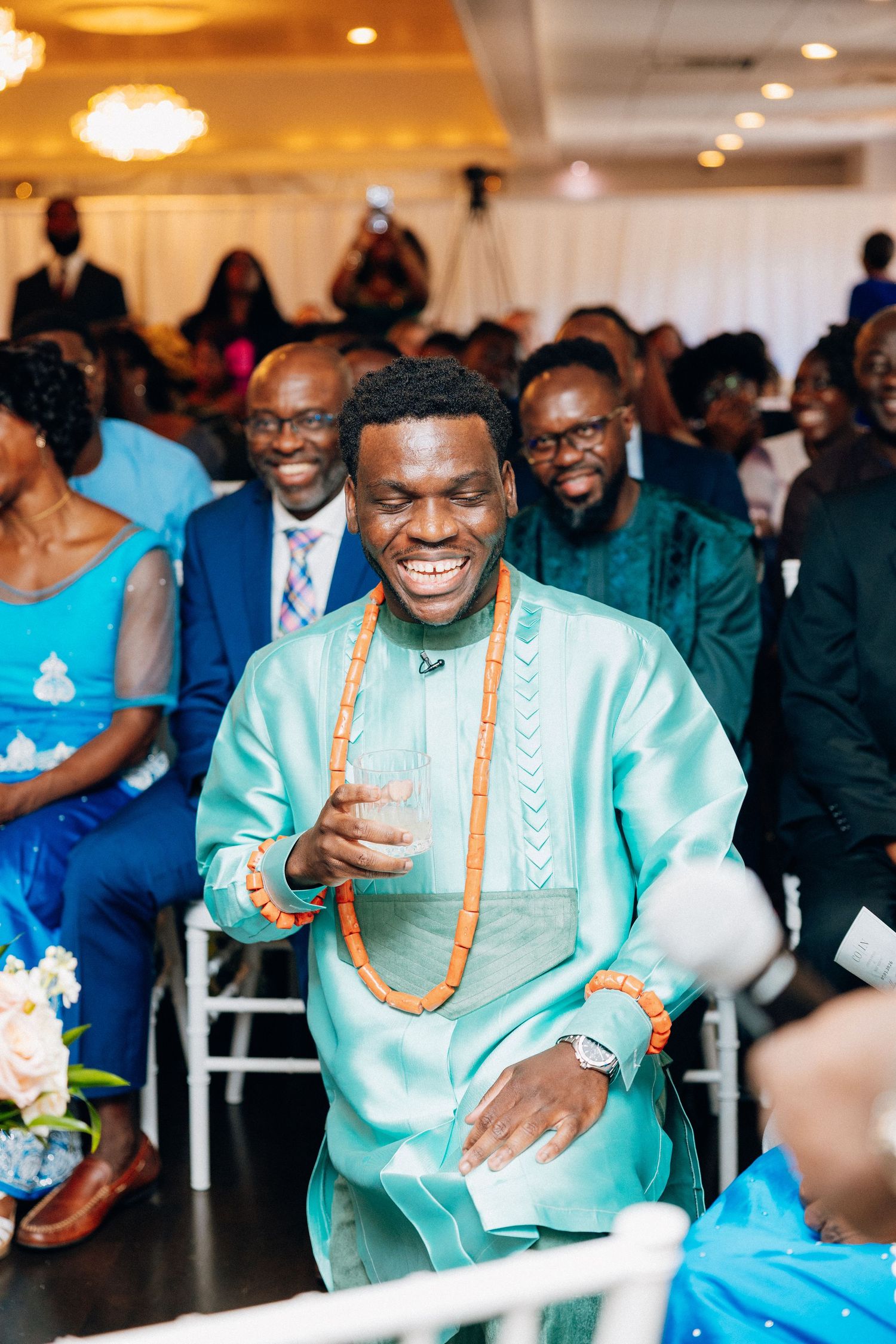 Groom in teal African attire smiles joyfully wearing orange beaded necklace at wedding ceremony.
