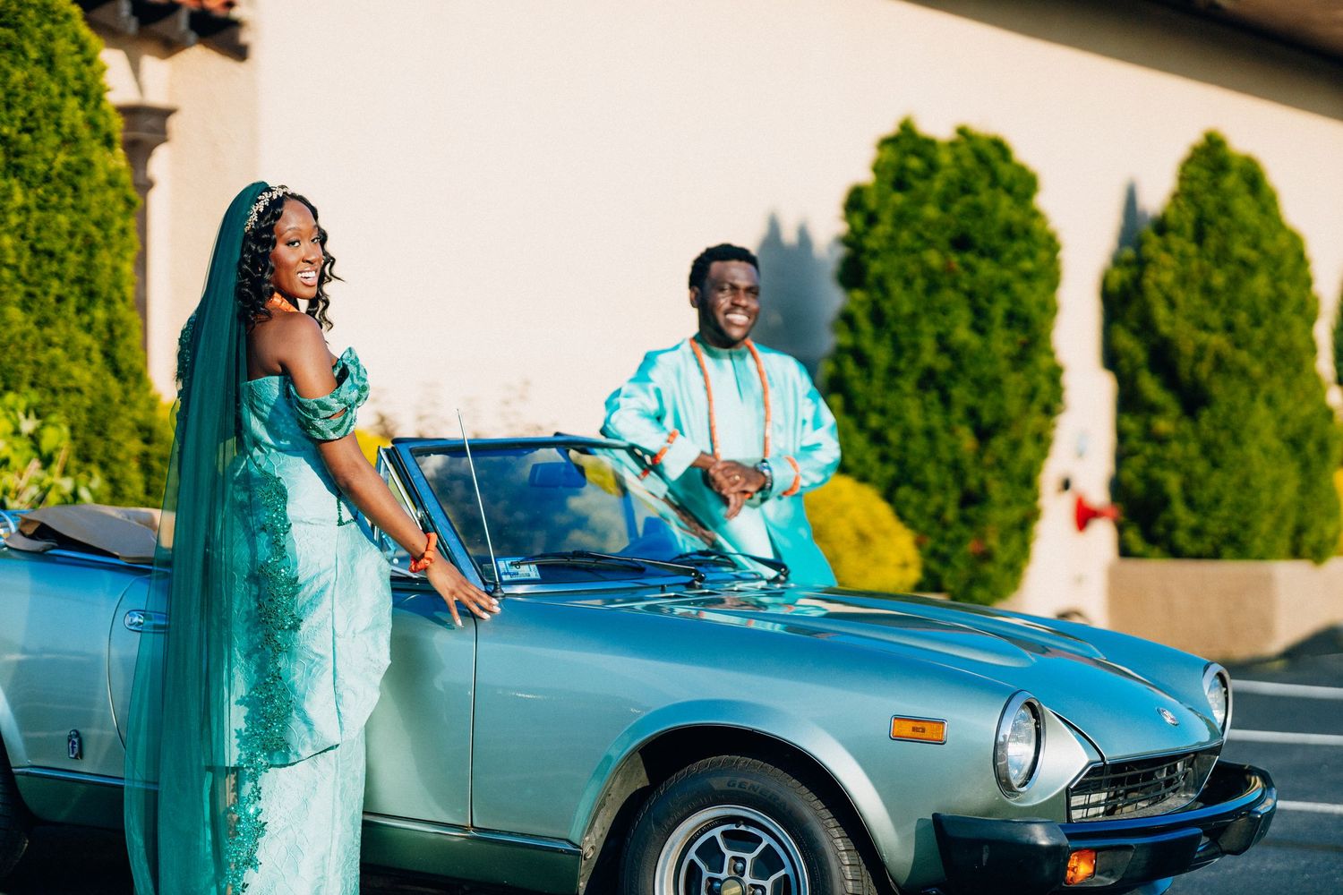 Bride and groom in teal attire pose beside vintage convertible.