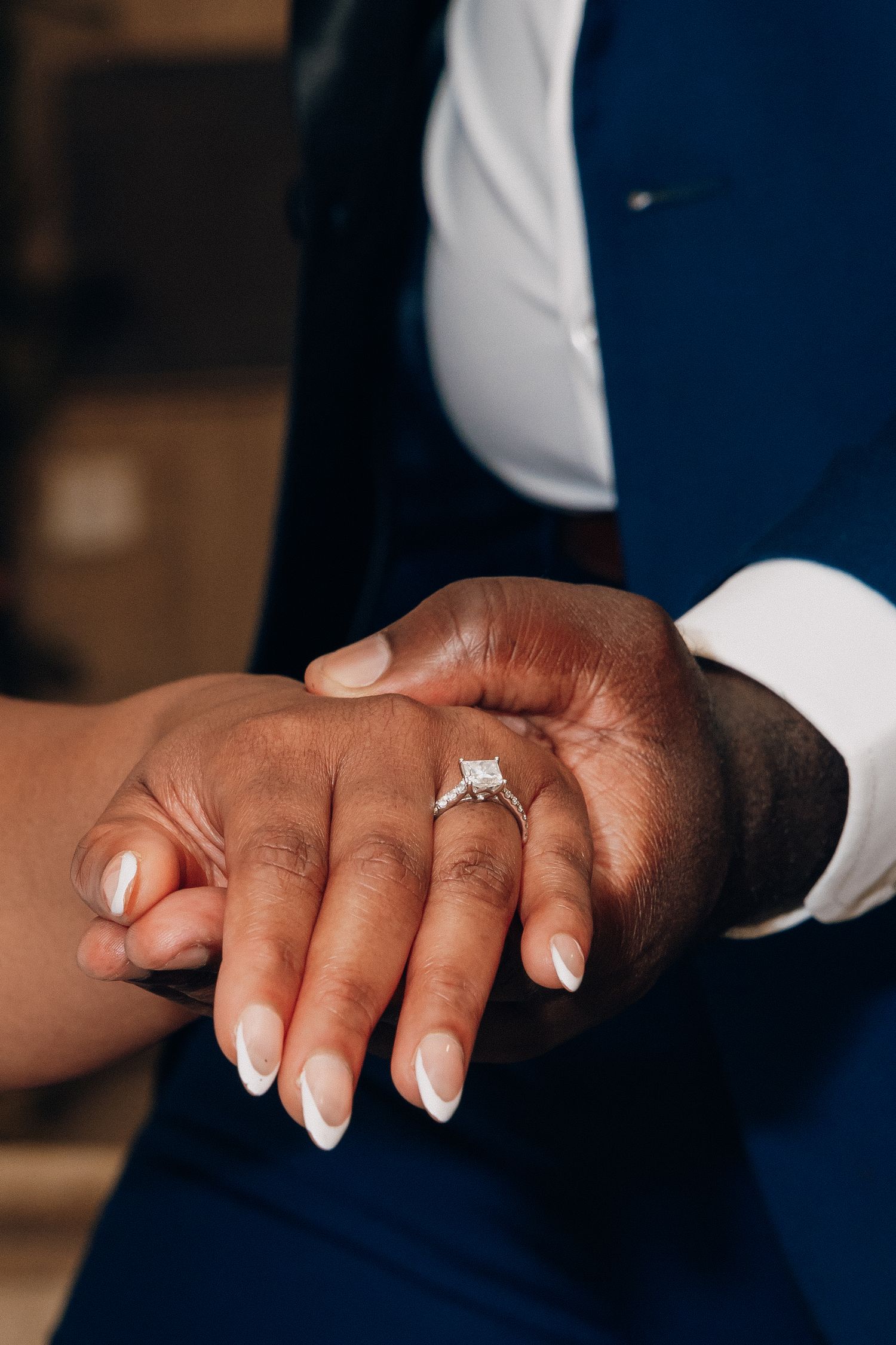 Close-up of an engagement ring on a woman's hand held by a man in a blue suit.