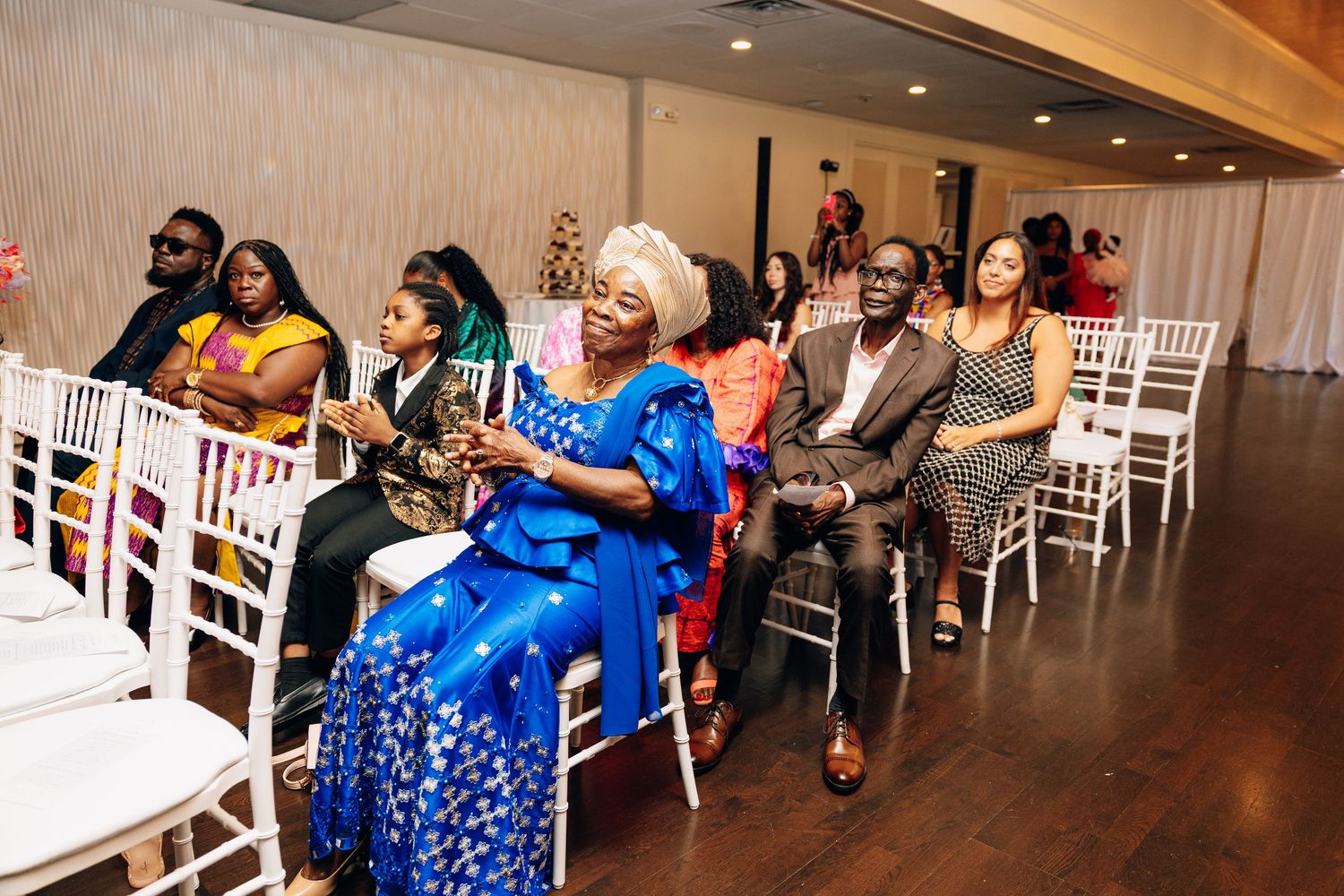 Guests in vibrant African attire sit in white chairs watching an elegant wedding ceremony indoors.