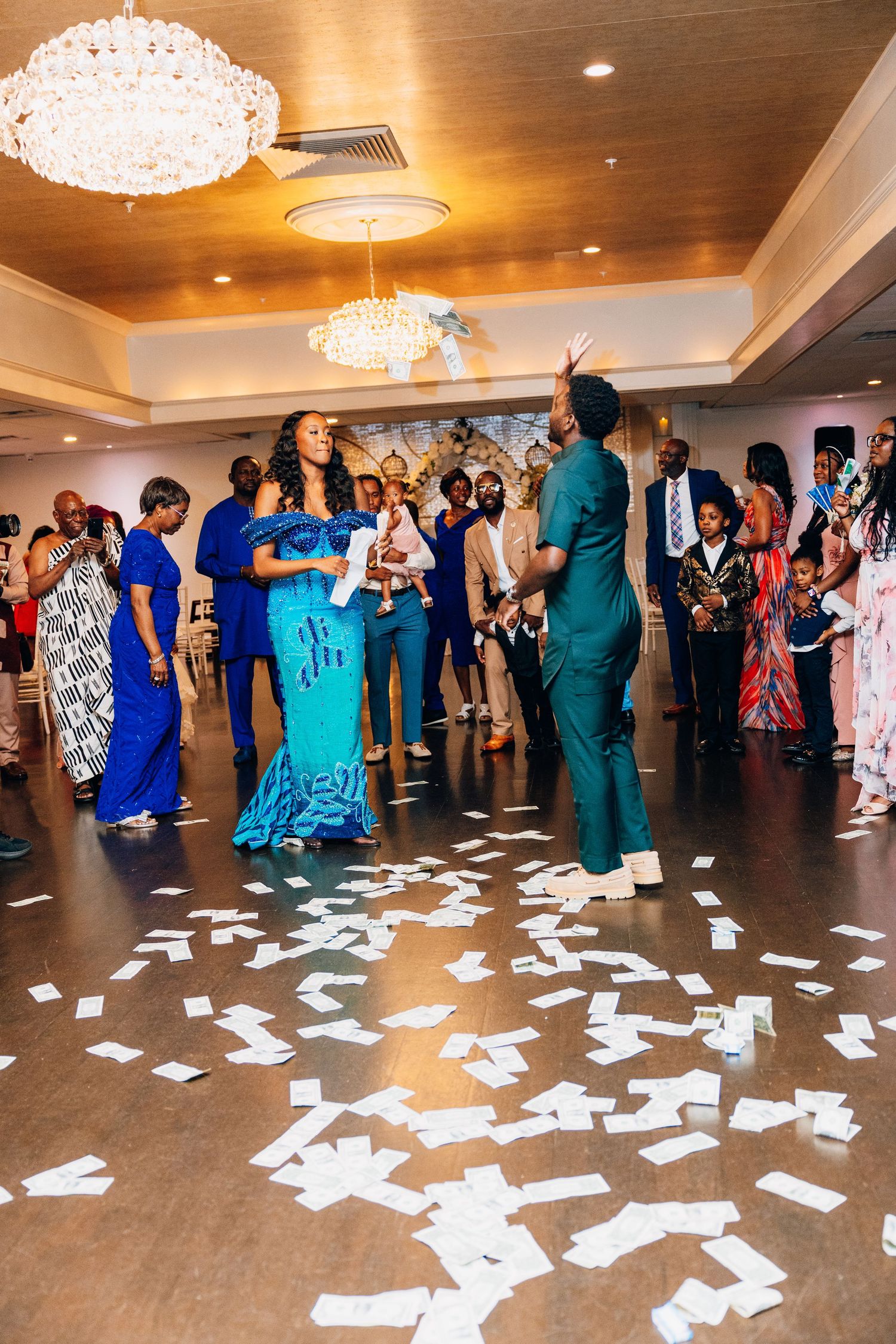 Wedding guests surround a couple on a dance floor covered in money during a lively reception celebration.