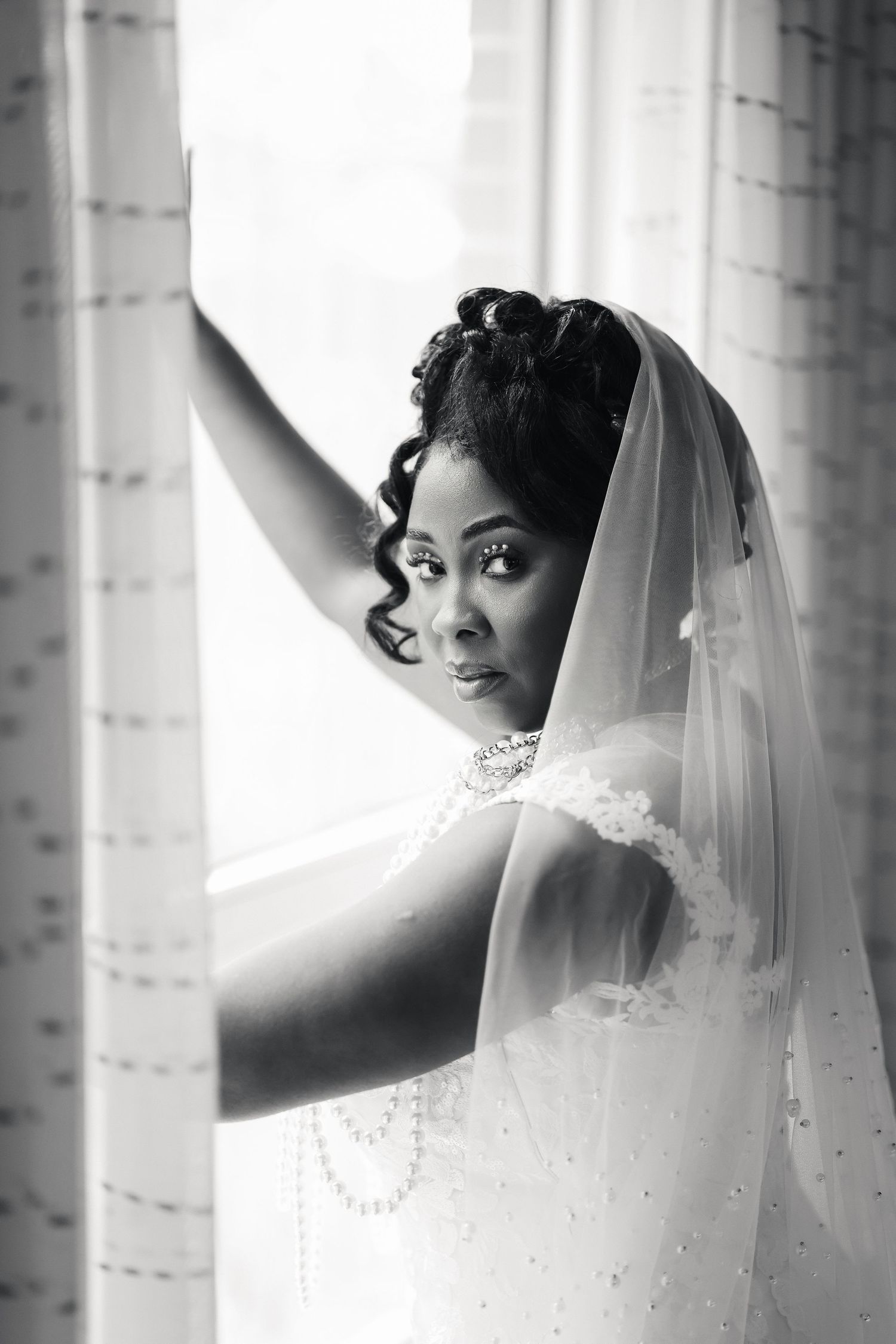 Black and white portrait of a bride in a veil and wedding dress standing by a window with curtains.