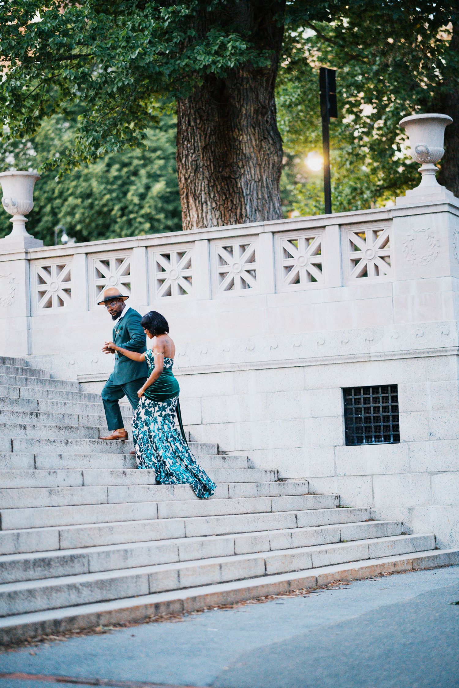 Couple in green formal attire descending white stone steps in sunlit park.