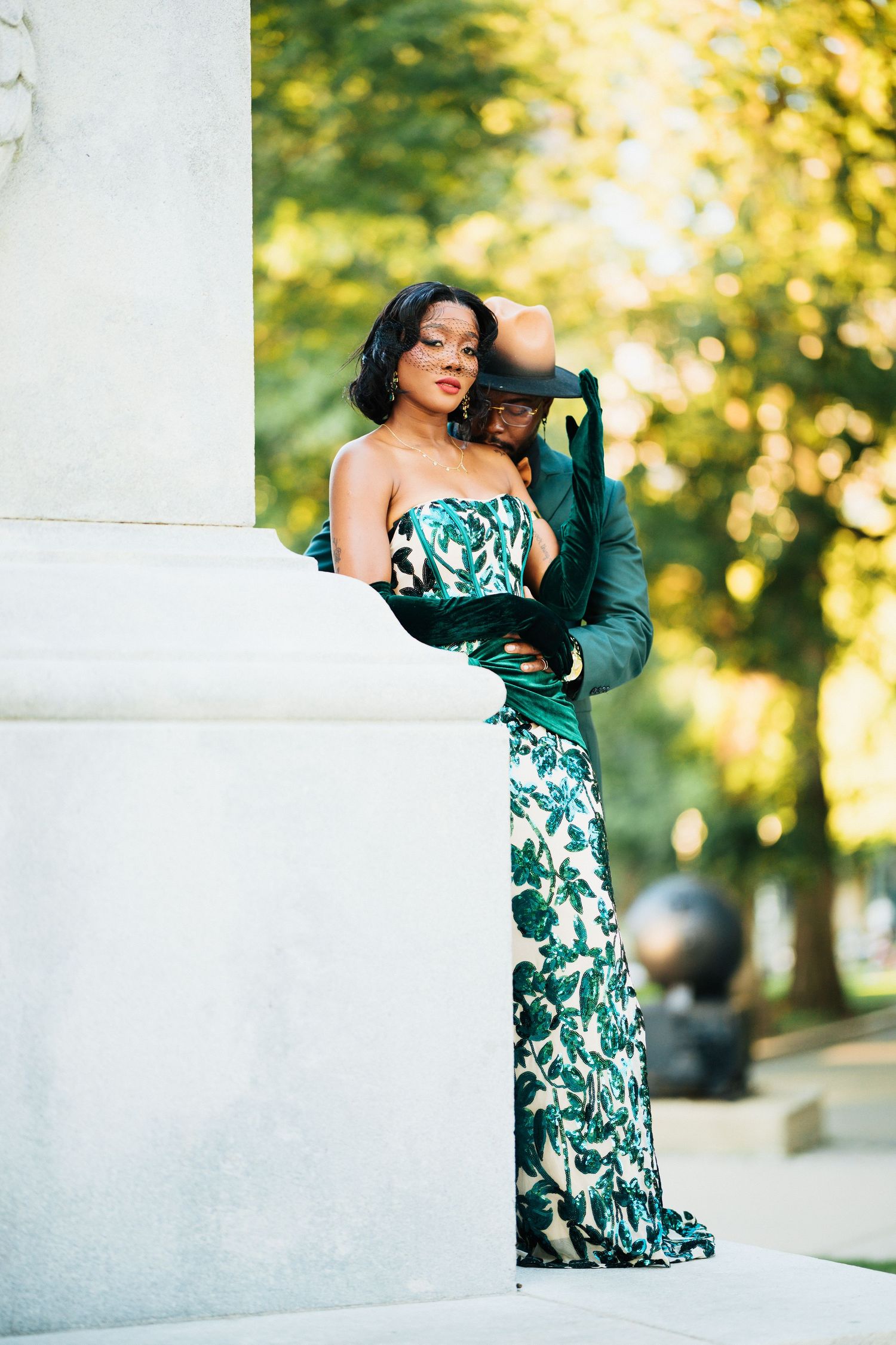 Couple in coordinating green outfits sharing intimate moment by a stone pillar in a sunlit park.