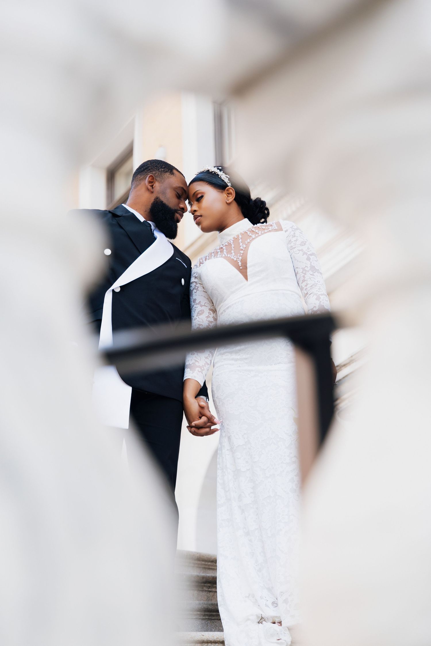 A Black bride and groom hold hands and gaze at each other on steps, she in lace gown, he in black tuxedo.
