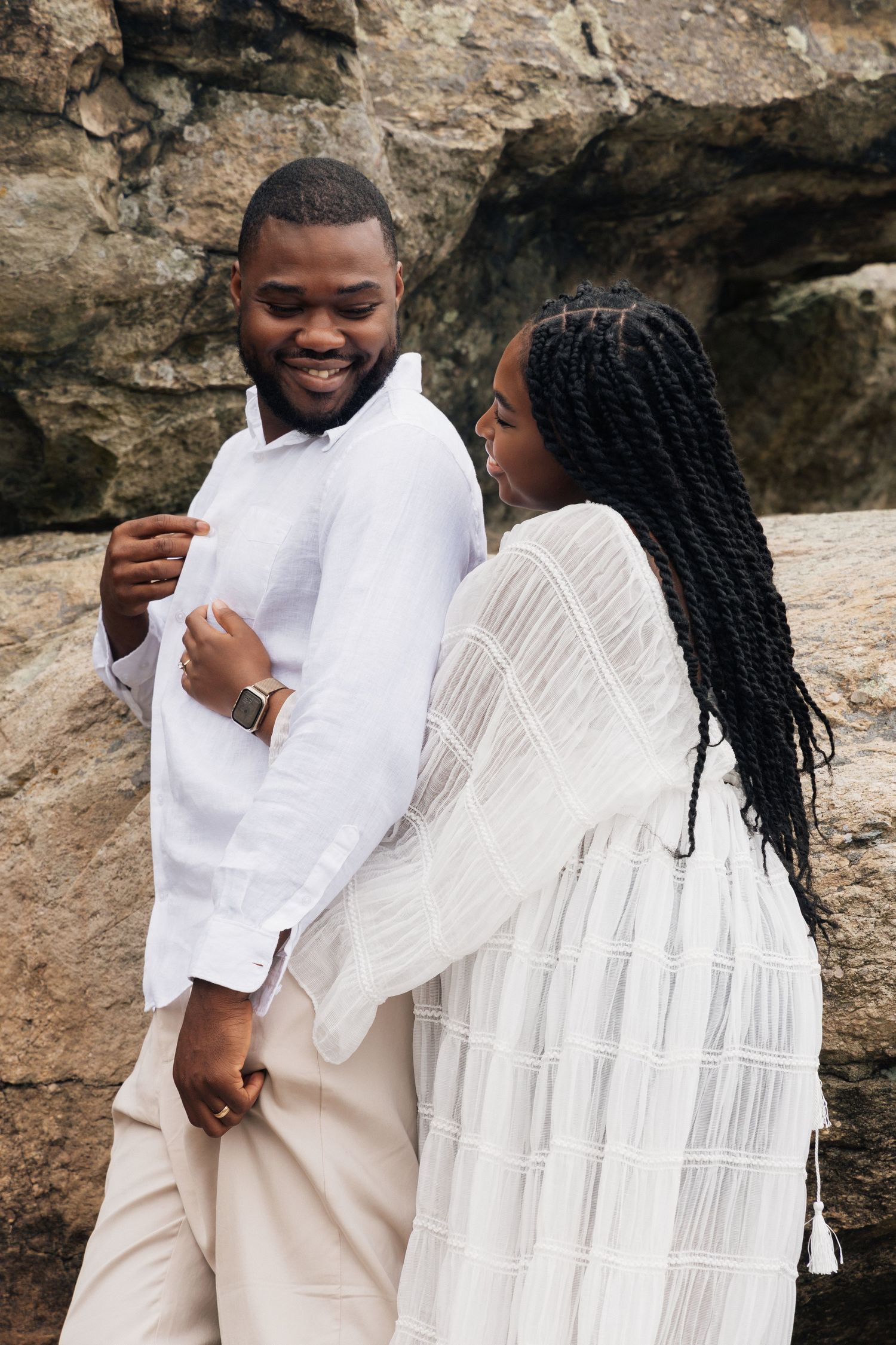 A smiling Black couple in coordinating white and beige outfits hold hands in front of a rocky backdrop.