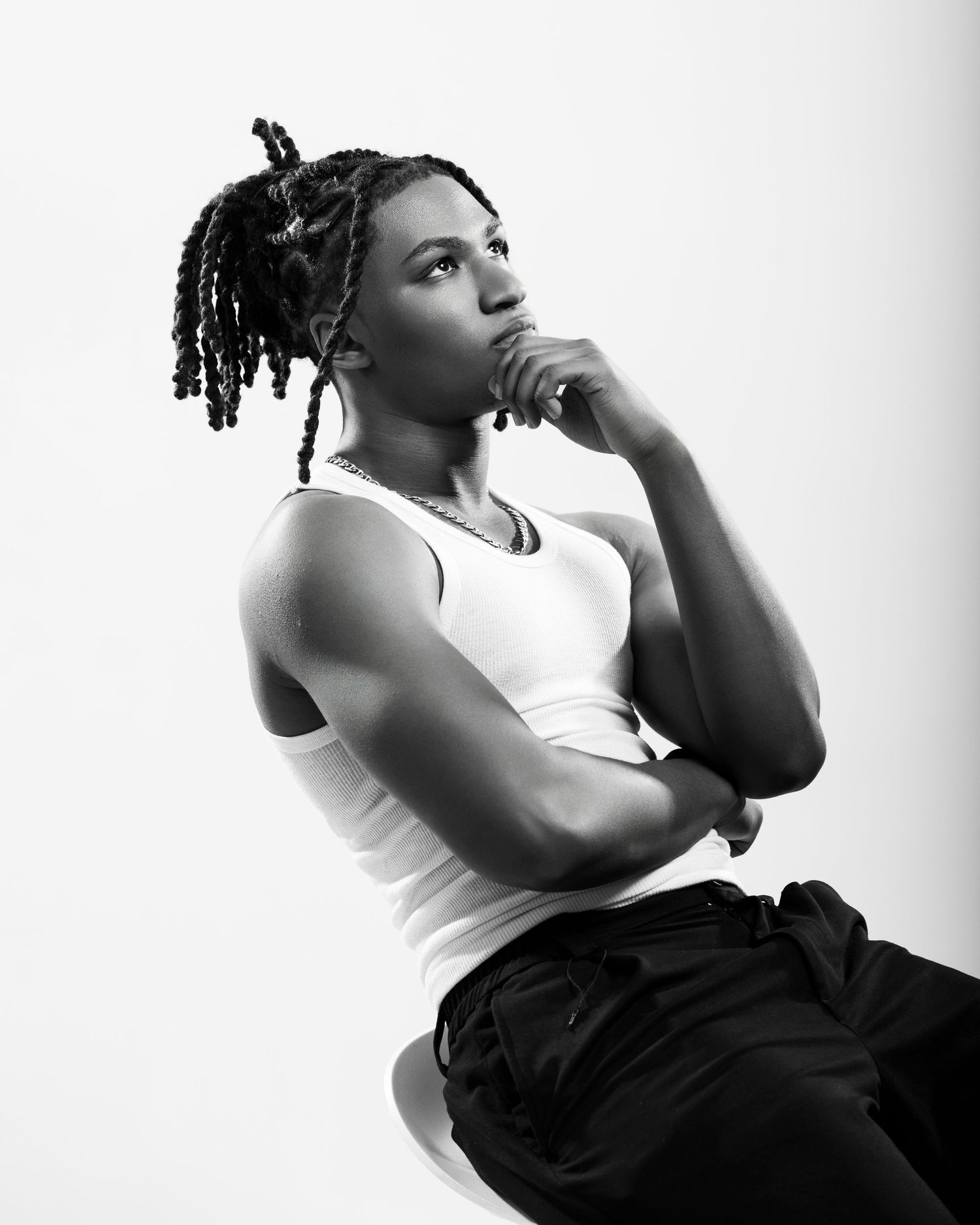 Black and white portrait of a young person with locs, wearing a white tank top, posing thoughtfully against a light background.