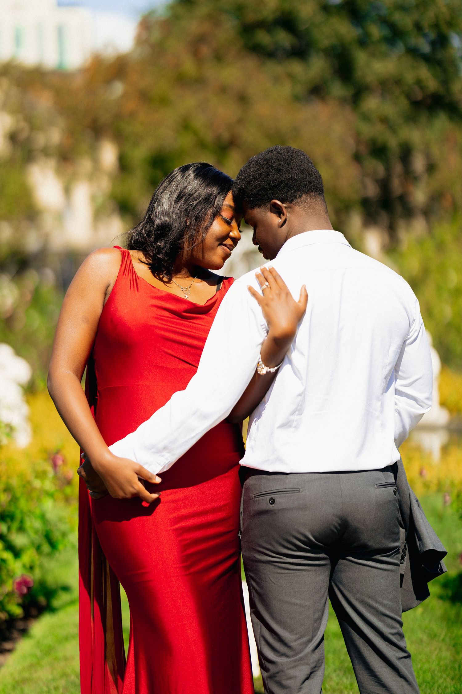 A couple in a garden, woman in red gown and man in white shirt share a romantic moment, touching foreheads.