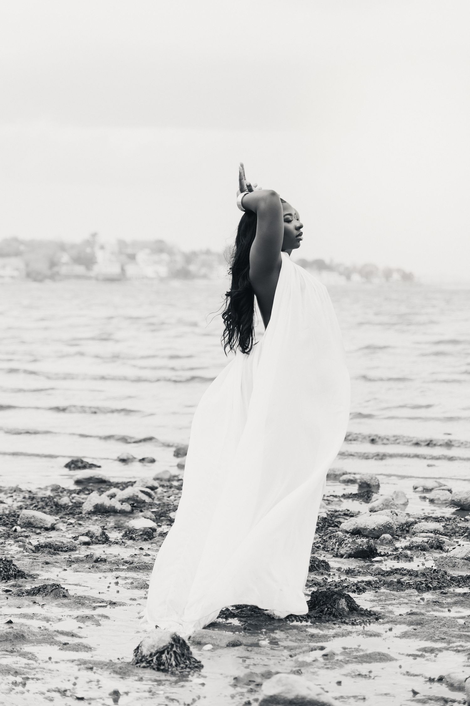 Black and white photo of a woman in flowing white fabric posing gracefully on a rocky shoreline with ocean in background.