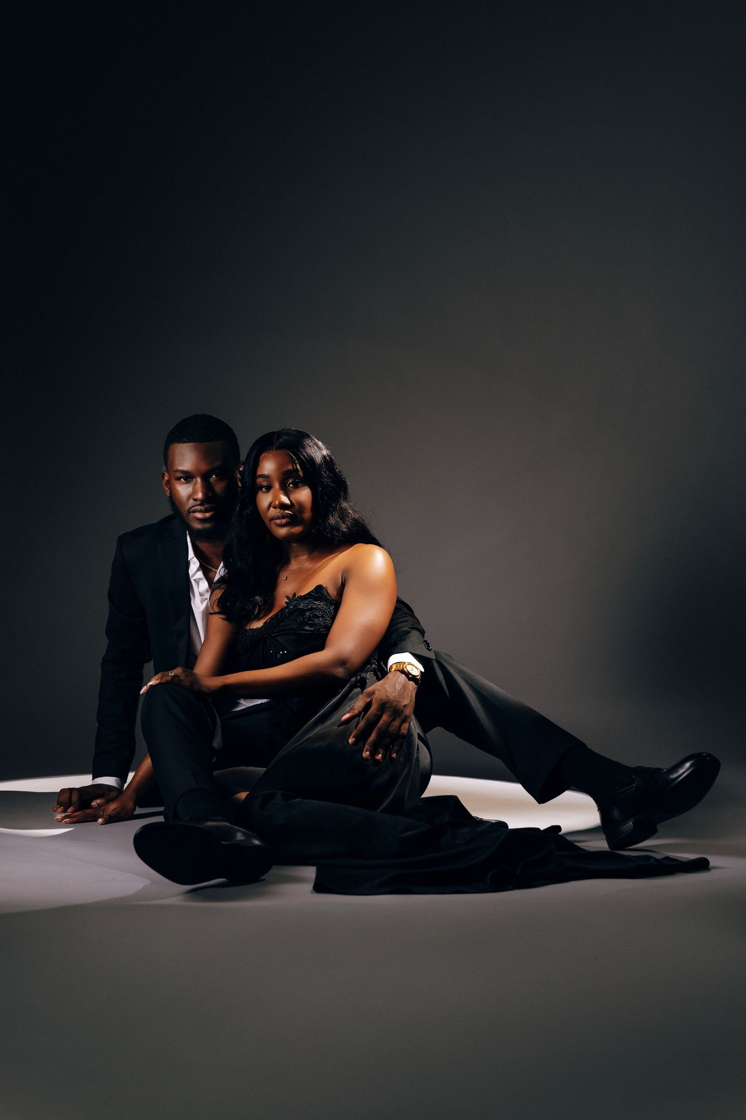 Elegant Black couple in formal attire sitting together on studio floor, woman leaning on man in dark romantic portrait.
