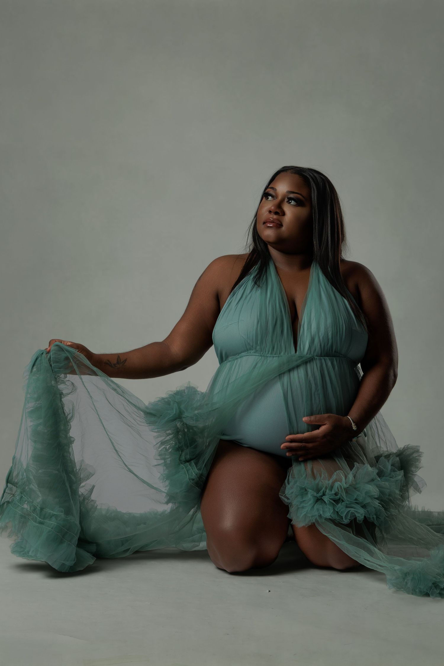 Pregnant woman in teal ruffle tulle gown kneeling gracefully in studio maternity photoshoot.
