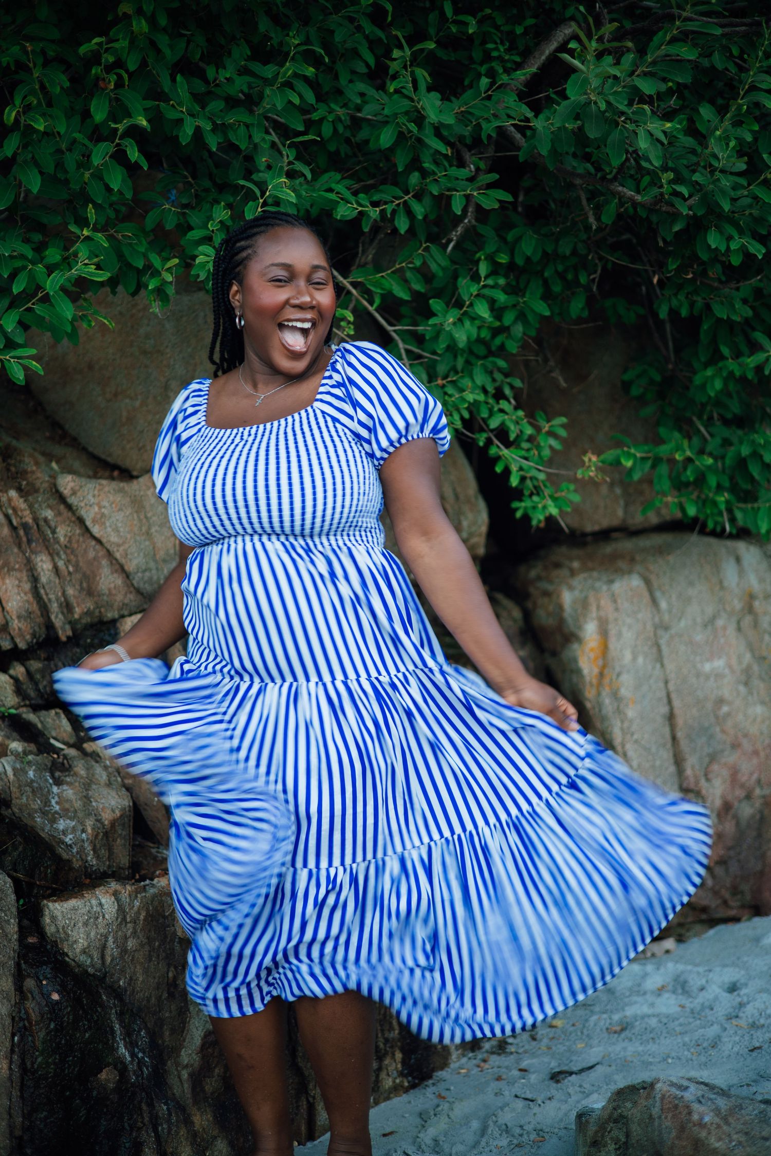 A joyful Black woman twirls in a blue and white striped dress outdoors near rocks and green foliage.