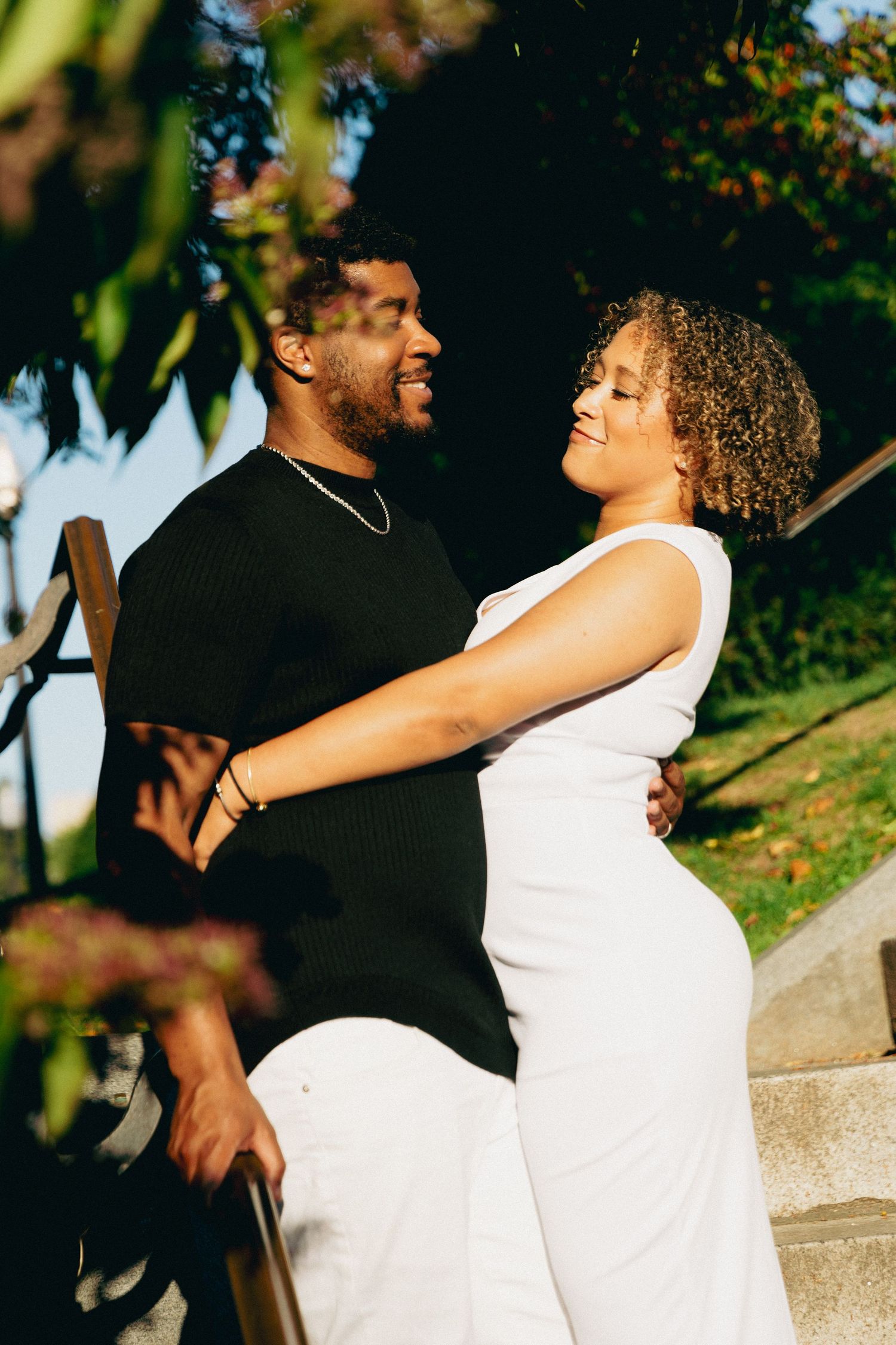 A smiling couple embraces outdoors on steps, woman in white dress and man in black shirt, surrounded by lush greenery.