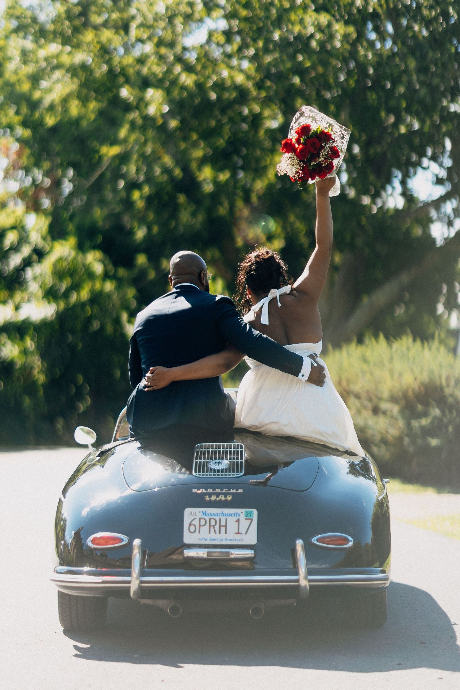 Bride raising red bouquet while sitting with groom on back of vintage black Porsche 356 convertible.