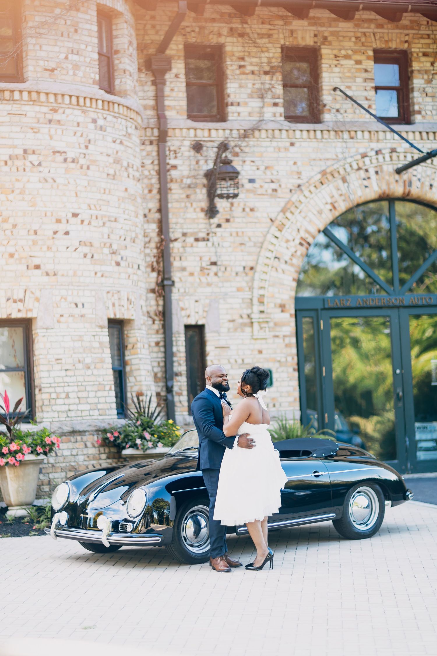 Bride and groom embracing beside a classic black Porsche convertible outside a stone venue.