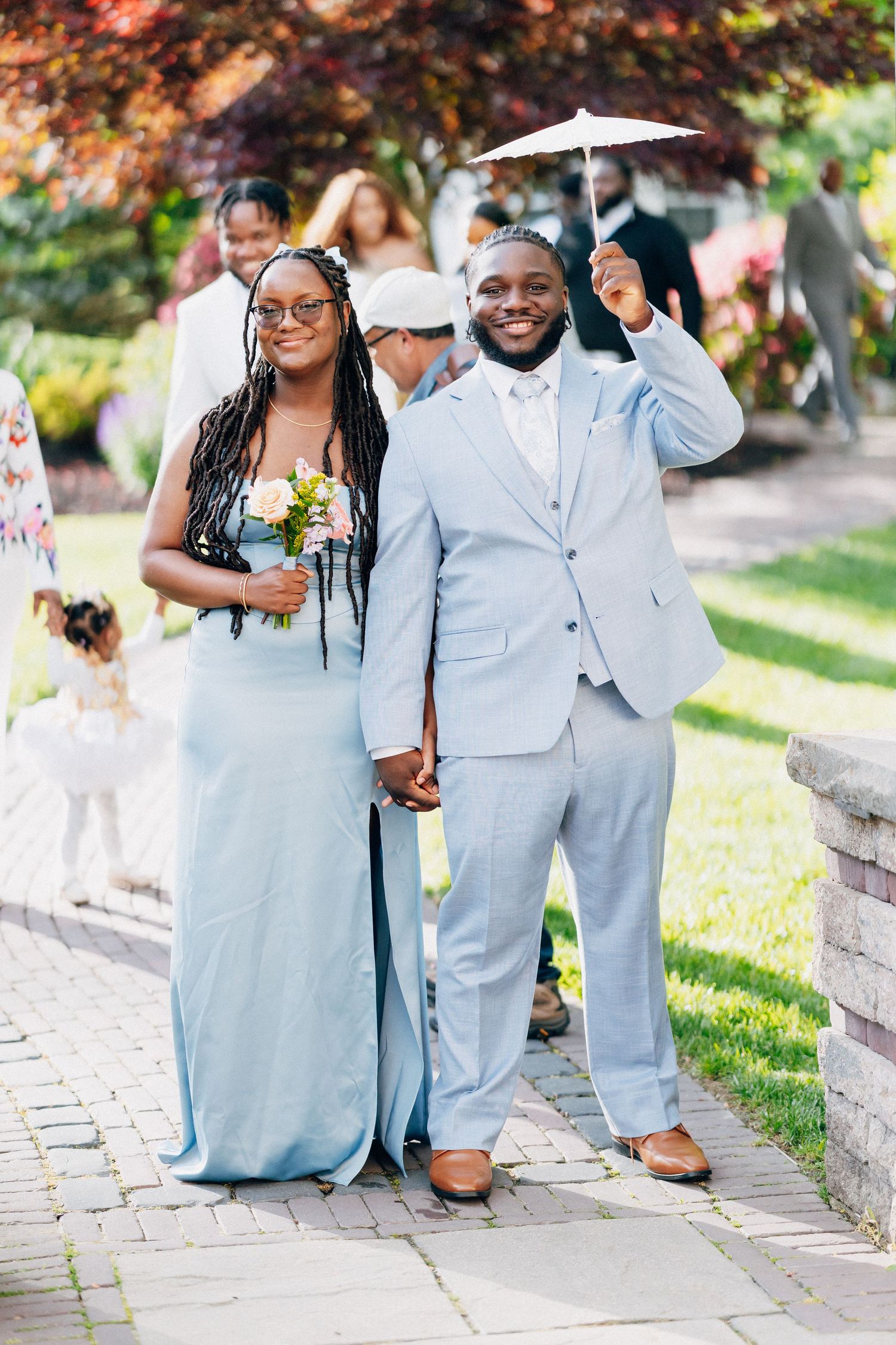 Bridesmaid in blue dress and groomsman in light blue suit smile holding parasol outdoors.