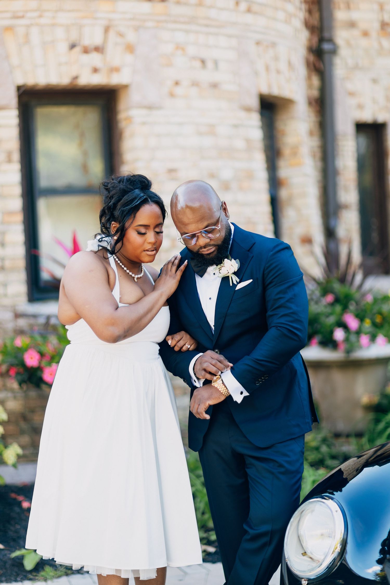 Bride and groom in wedding attire stand beside a vintage car outside a brick building with colorful flowers.