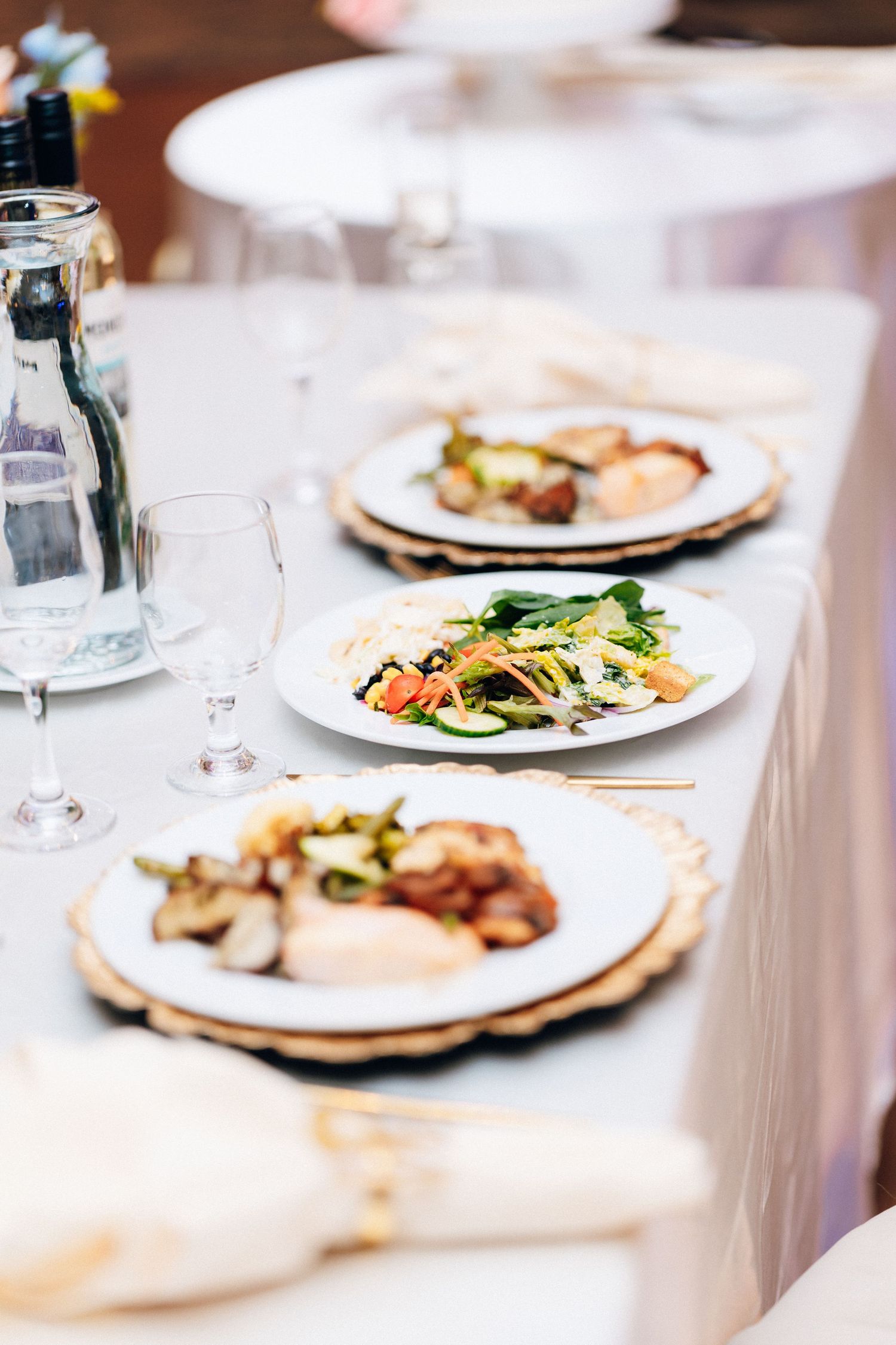 Elegant banquet table set with white plates of gourmet food, wine glasses, and gold chargers for a formal event.