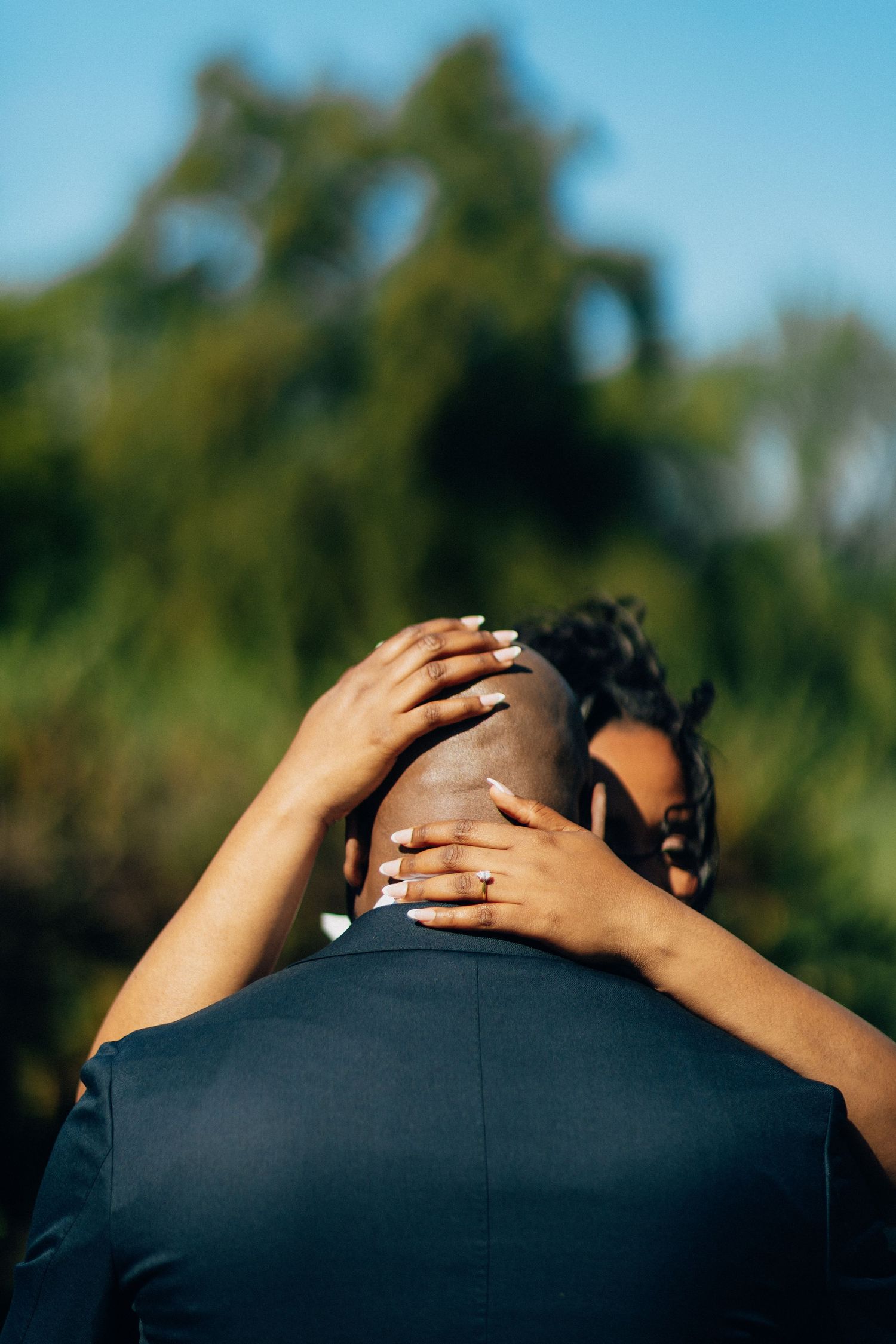 Bride placing hands on grooms shaved head in a tender embrace with green trees in background.