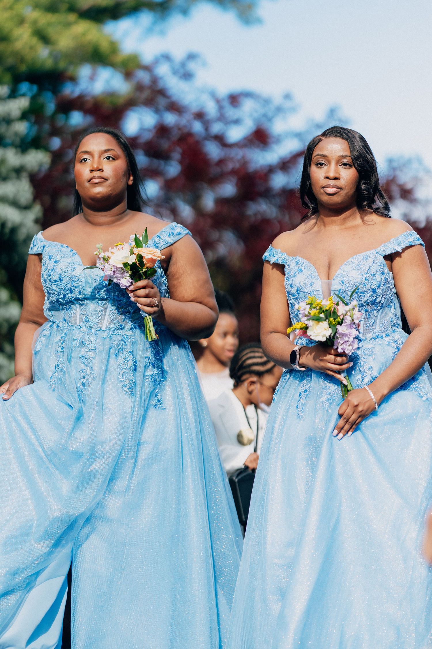 Two bridesmaids in matching light blue off-shoulder gowns holding colorful bouquets at an outdoor wedding ceremony.