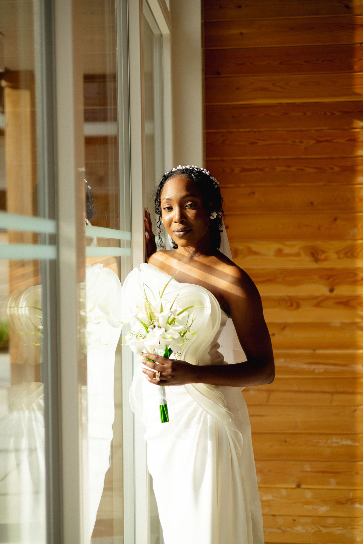 A bride in a white off-shoulder gown holds a calla lily bouquet while standing by a sunlit wooden wall.