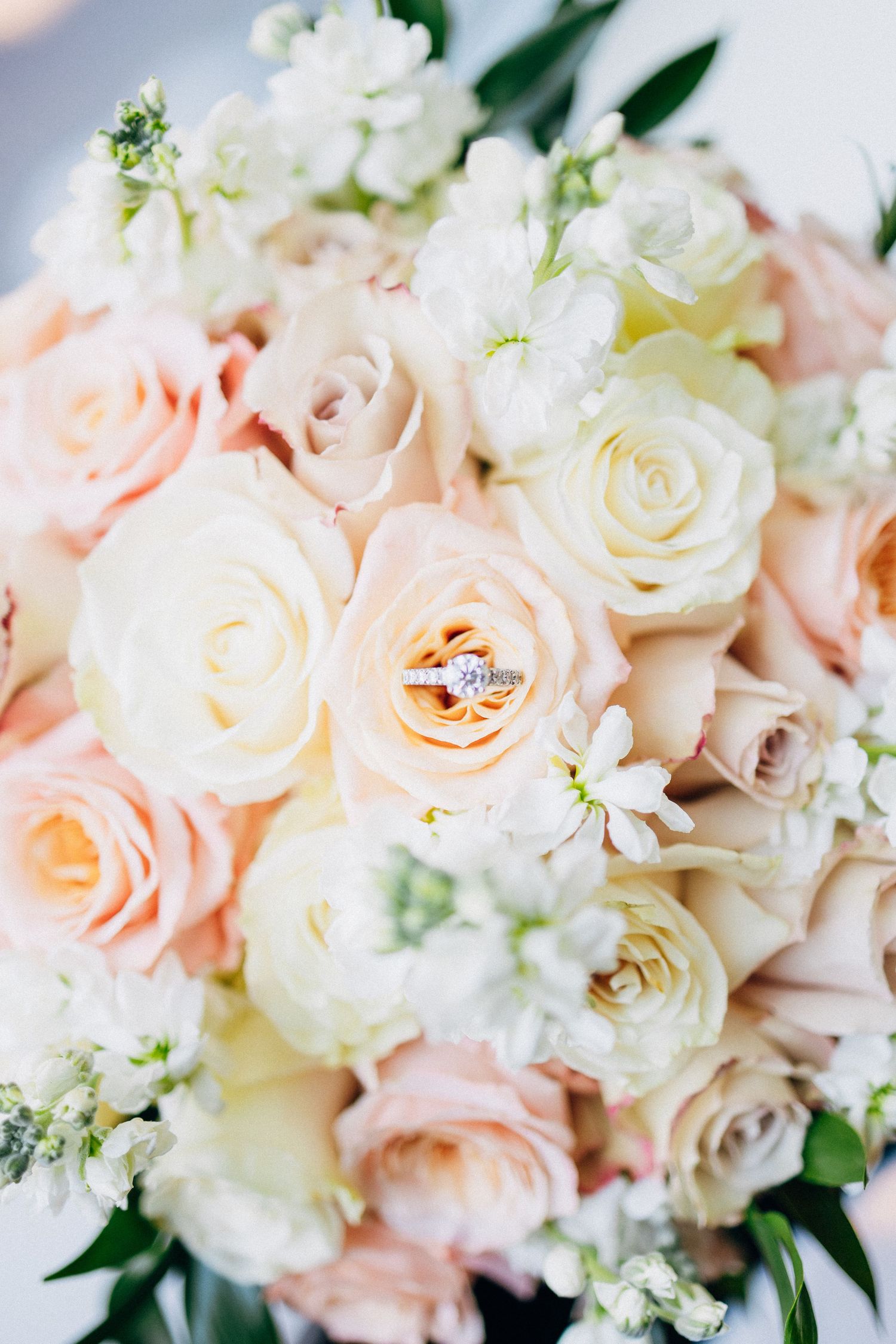 Elegant bridal bouquet of blush and white roses with a diamond engagement ring nestled among the blooms.