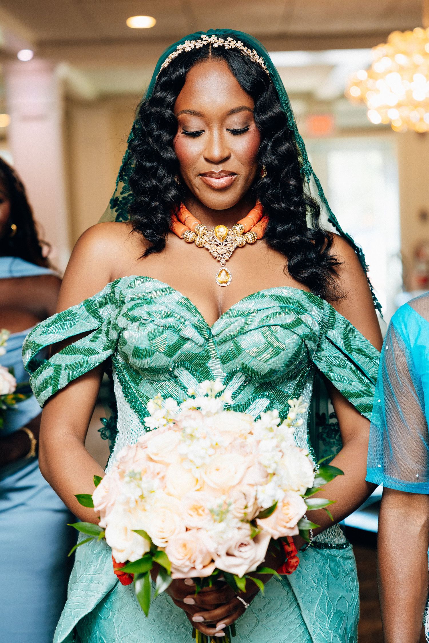 Bride in teal African lace gown with orange jewelry and veil holds white rose bouquet at traditional wedding ceremony.
