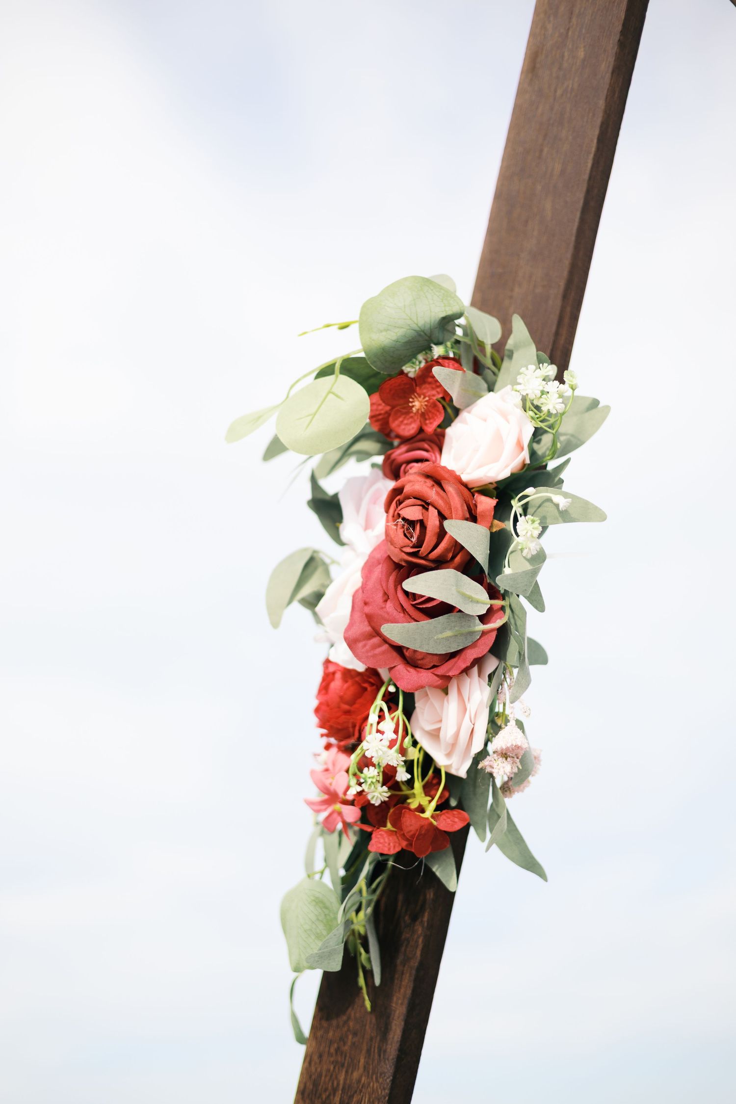 A wooden wedding arch adorned with red and pink roses and eucalyptus greenery against a cloudy sky.