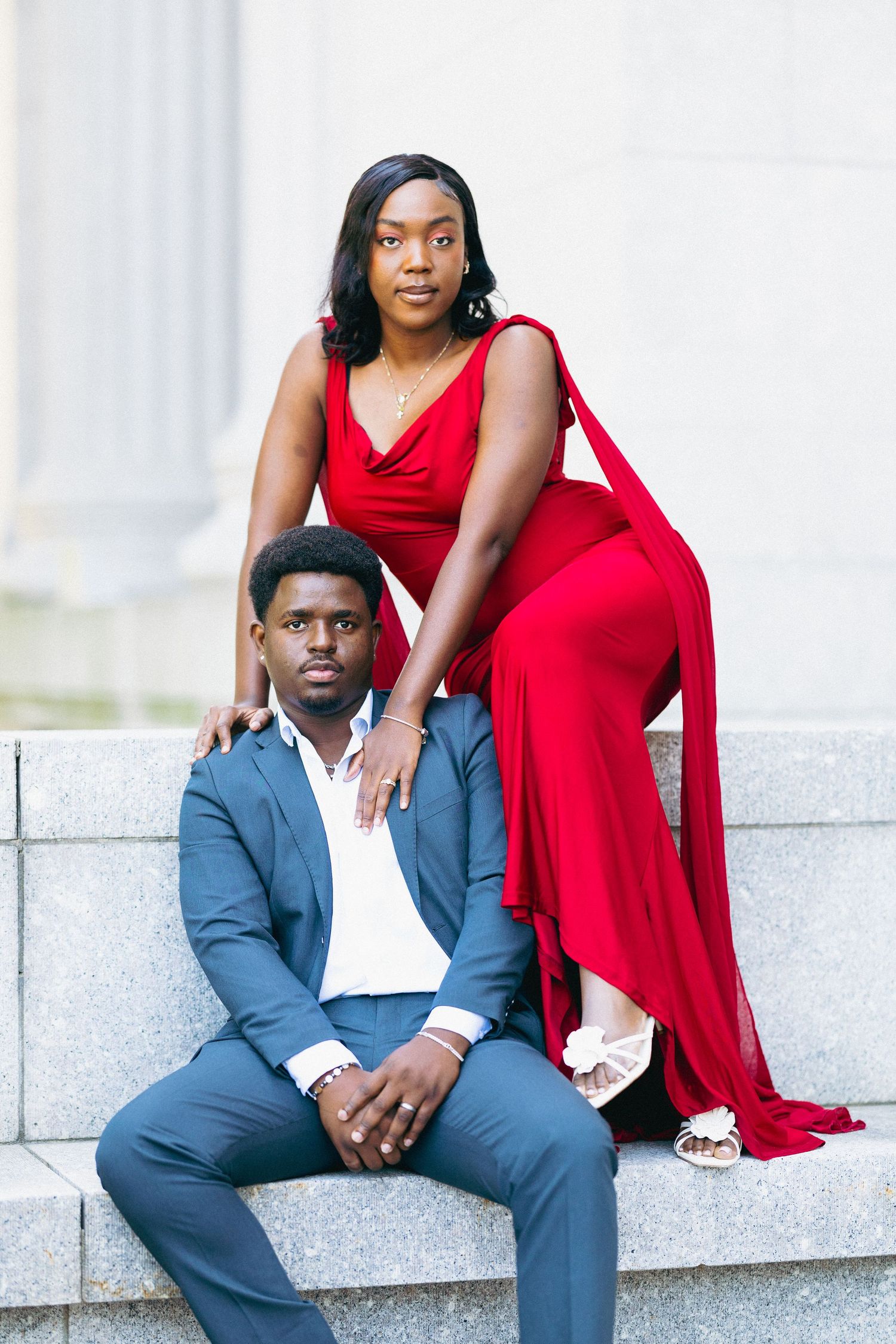 A couple poses on stone steps, woman in red gown stands behind man in blue suit for a formal portrait.