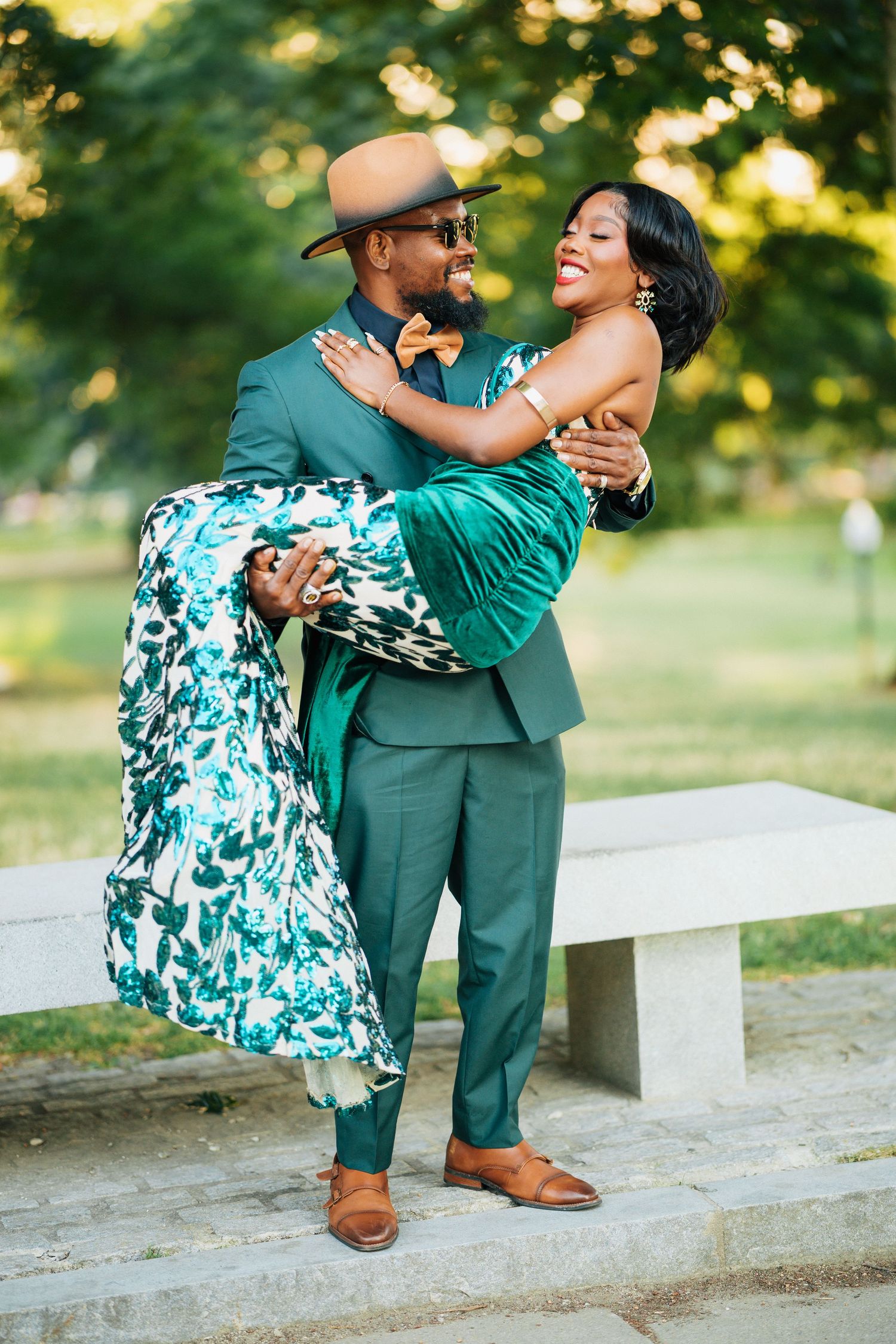 Stylish man in green suit lifting smiling woman in floral dress in a park.