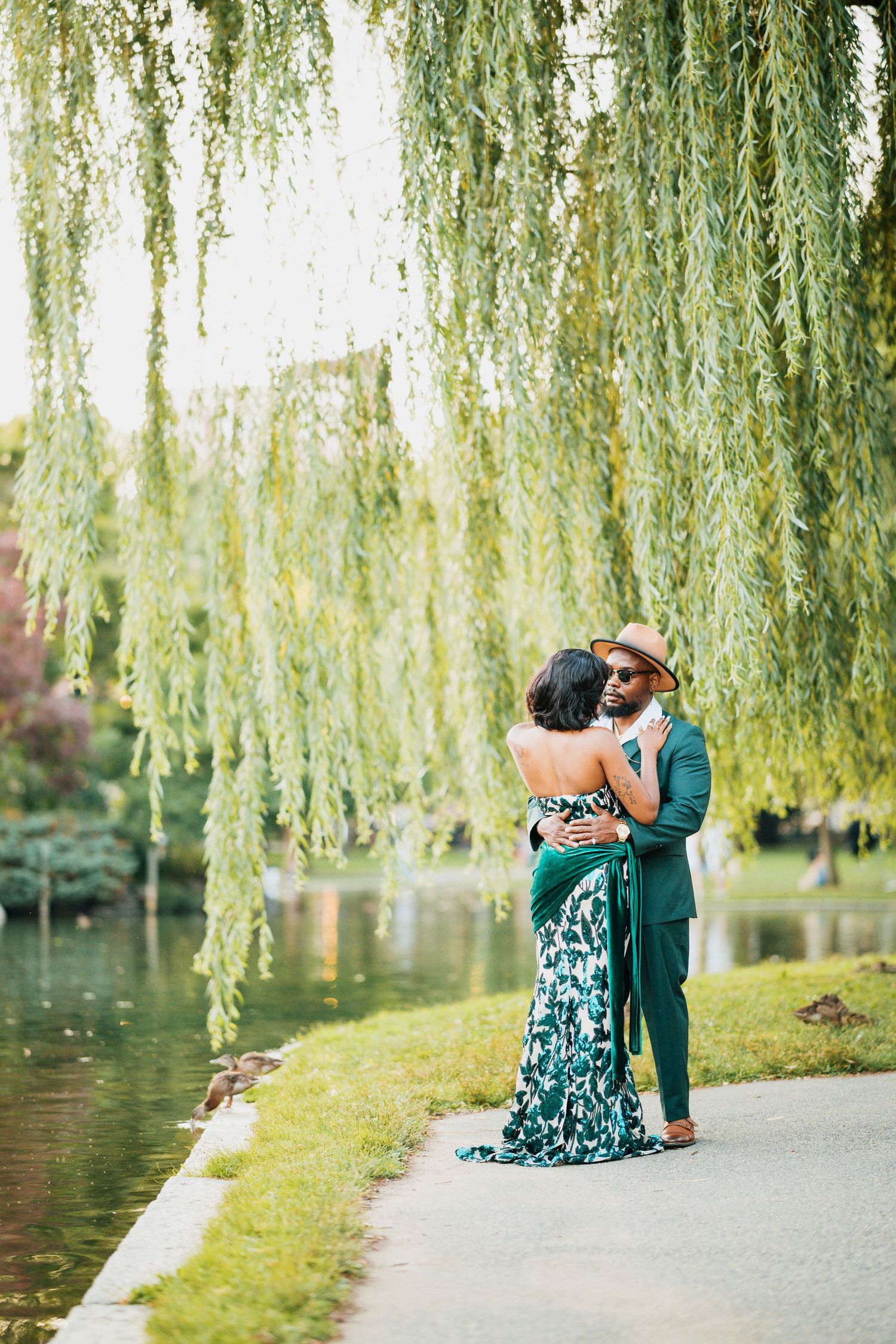 Couple embracing under weeping willow tree by a serene pond in a park.