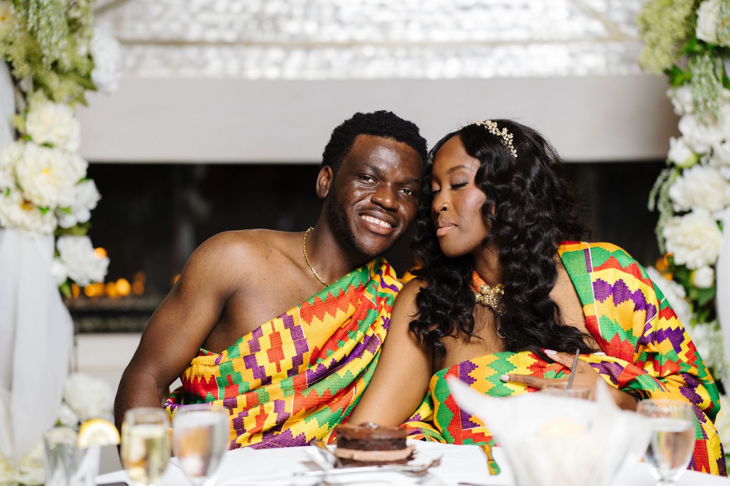 A smiling couple in matching Kente cloth outfits share a tender moment at their traditional African wedding.