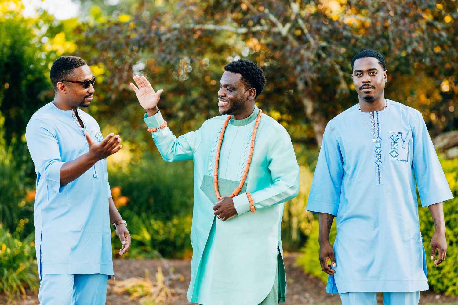 Three men in traditional African attire smile and high-five outdoors in a lush green setting.