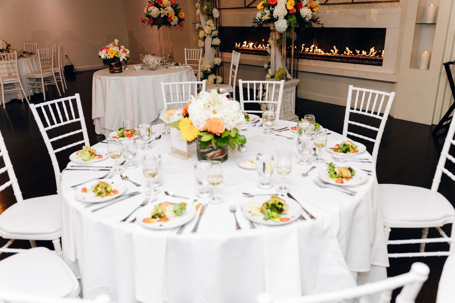 Elegant wedding reception table with white linens, Chiavari chairs, floral centerpiece, and a modern fireplace backdrop.