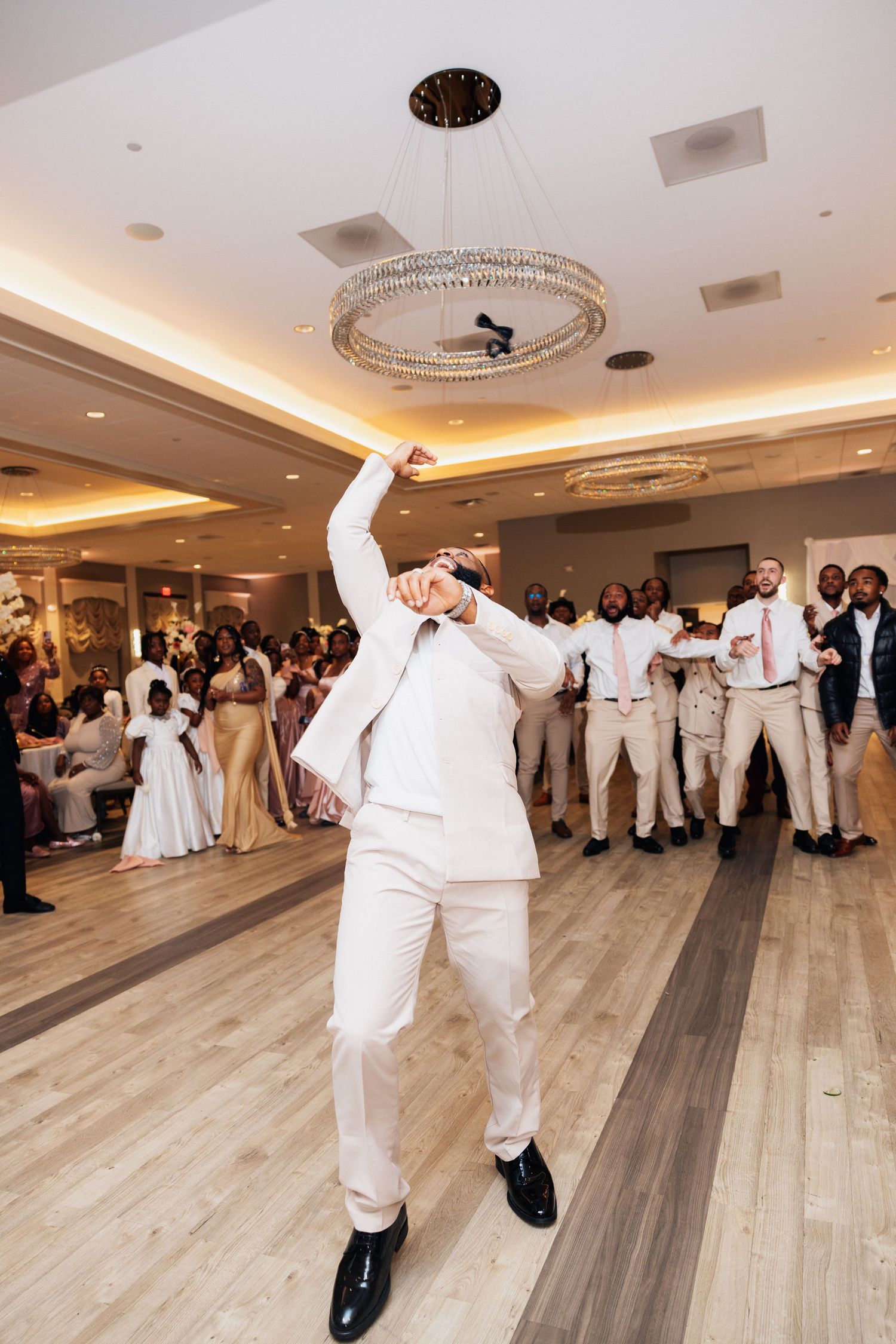 Groom in white suit dancing energetically at wedding reception with groomsmen performing choreographed dance behind him.