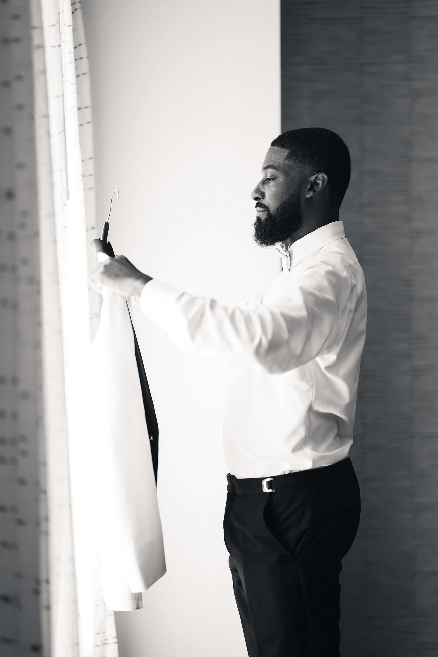Black and white photo of a bearded man in white shirt holding a suit jacket near a bright window.