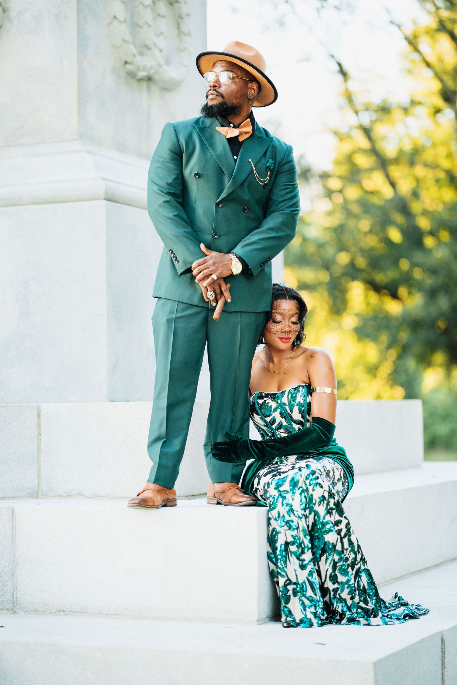 Elegant Black couple posing on white marble steps, man in green suit and hat, woman in floral green gown.