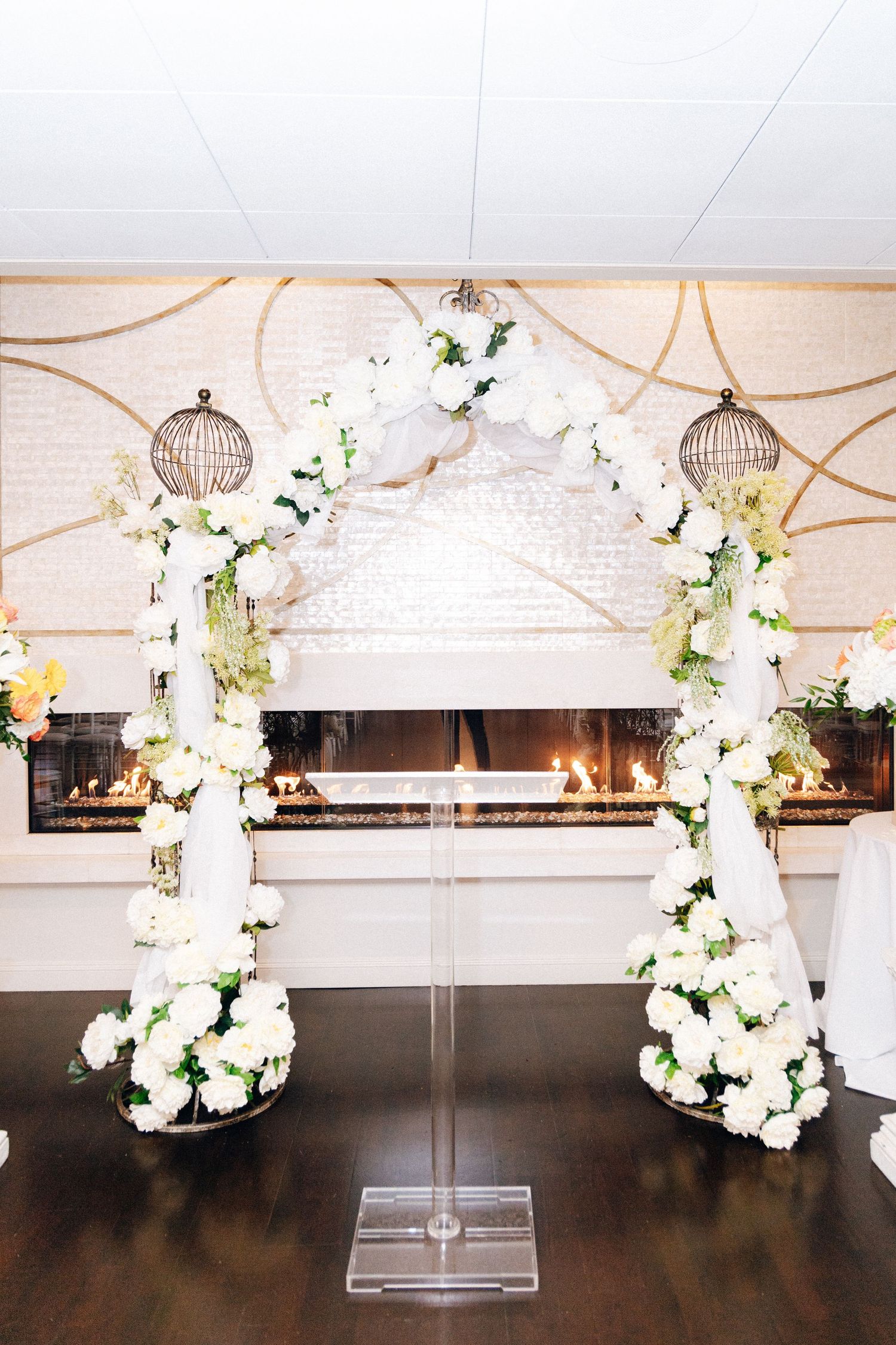 Elegant wedding arch adorned with white roses and greenery beside a lit fireplace.