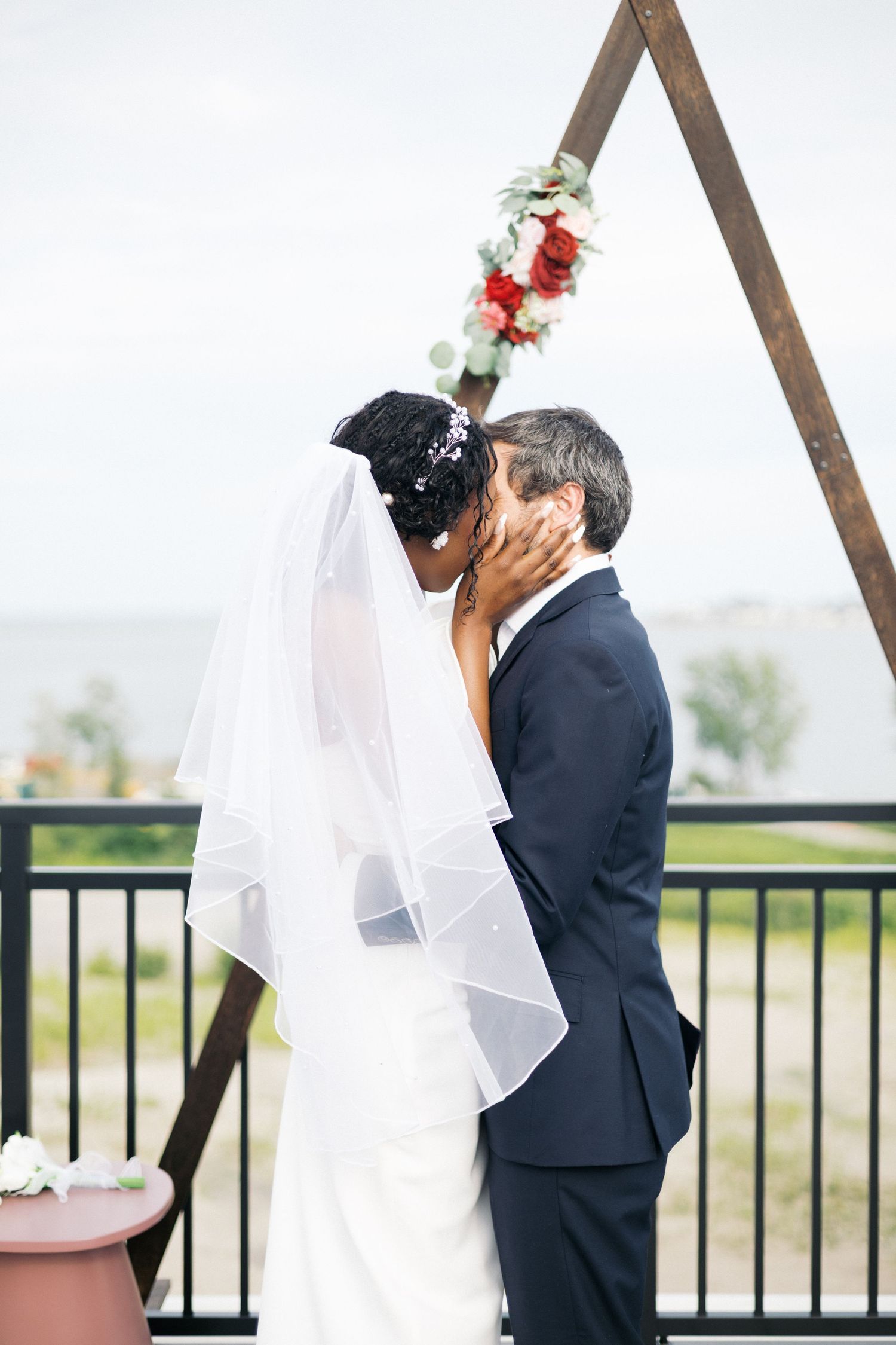 A bride in a white veil and groom in a navy suit share a romantic kiss at their outdoor wedding ceremony.