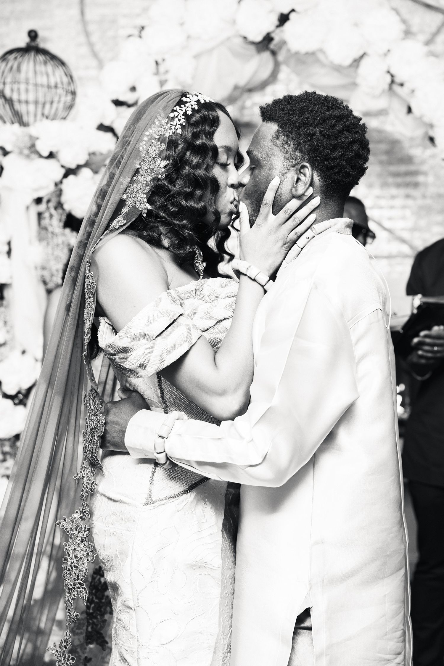 Black and white photo of bride and groom sharing first kiss at wedding altar with floral backdrop.
