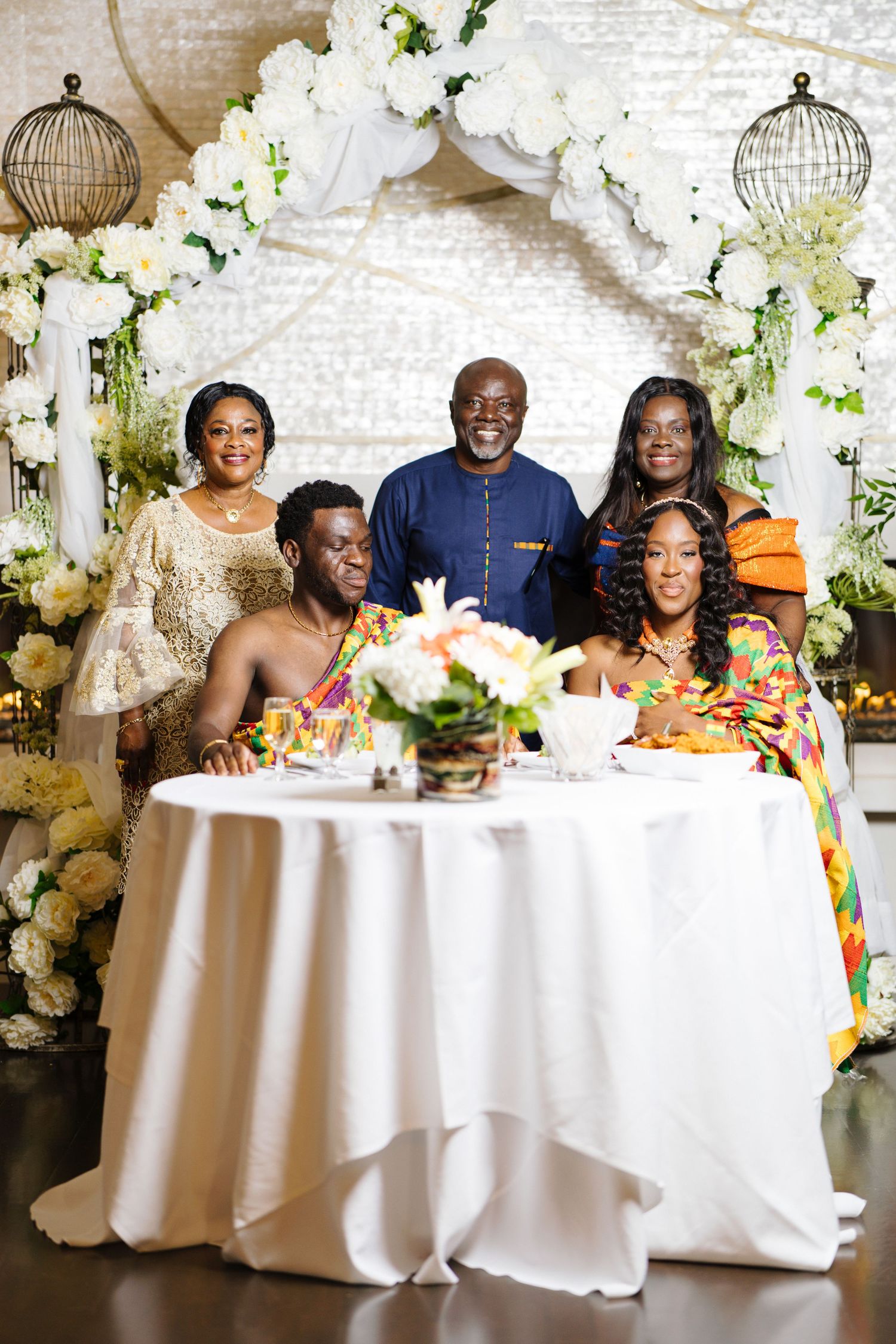 Family portrait of five people posing at a wedding sweetheart table with white floral arch backdrop.