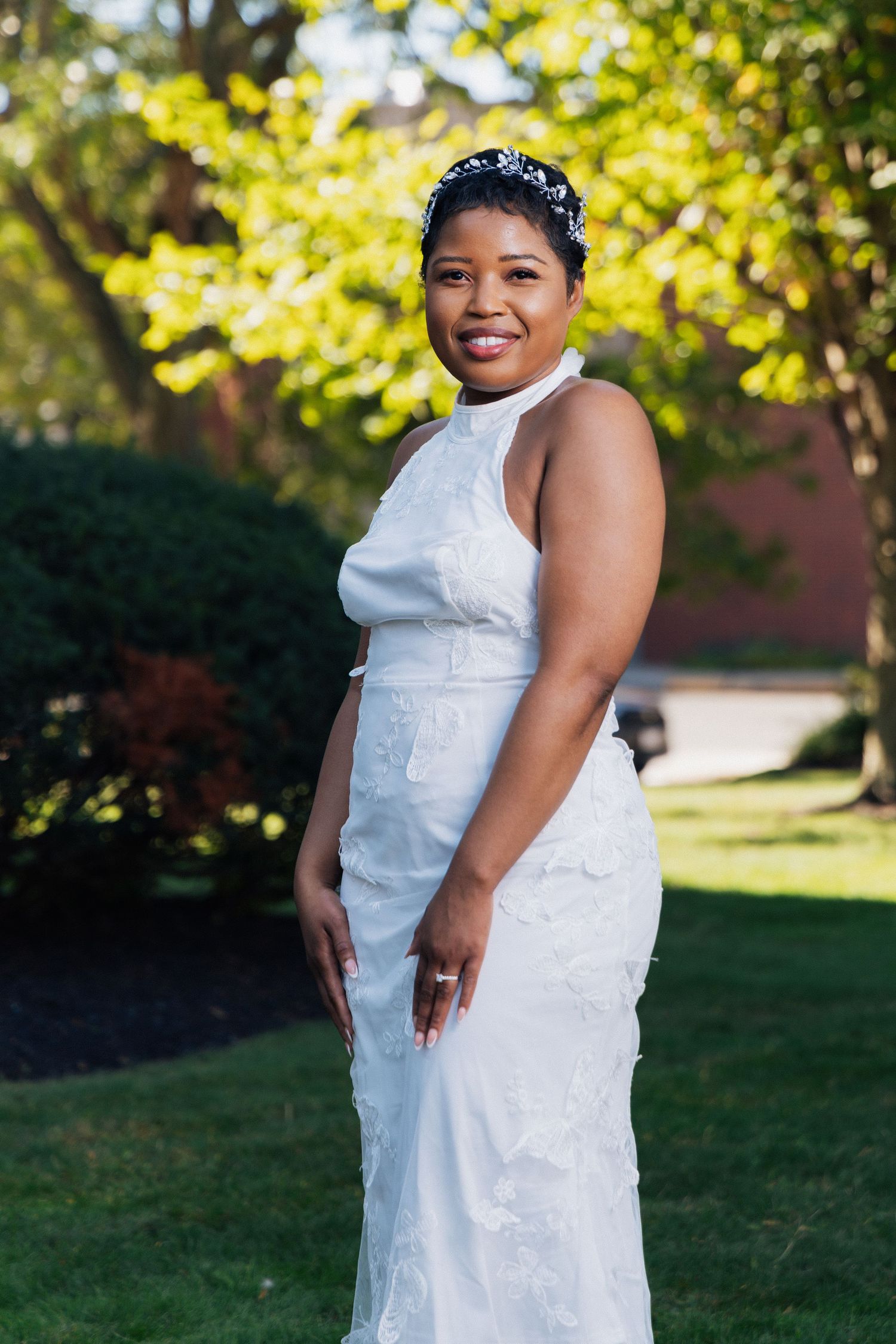 Elegant bride in white lace halter wedding gown with crystal headpiece smiling in sunny garden.